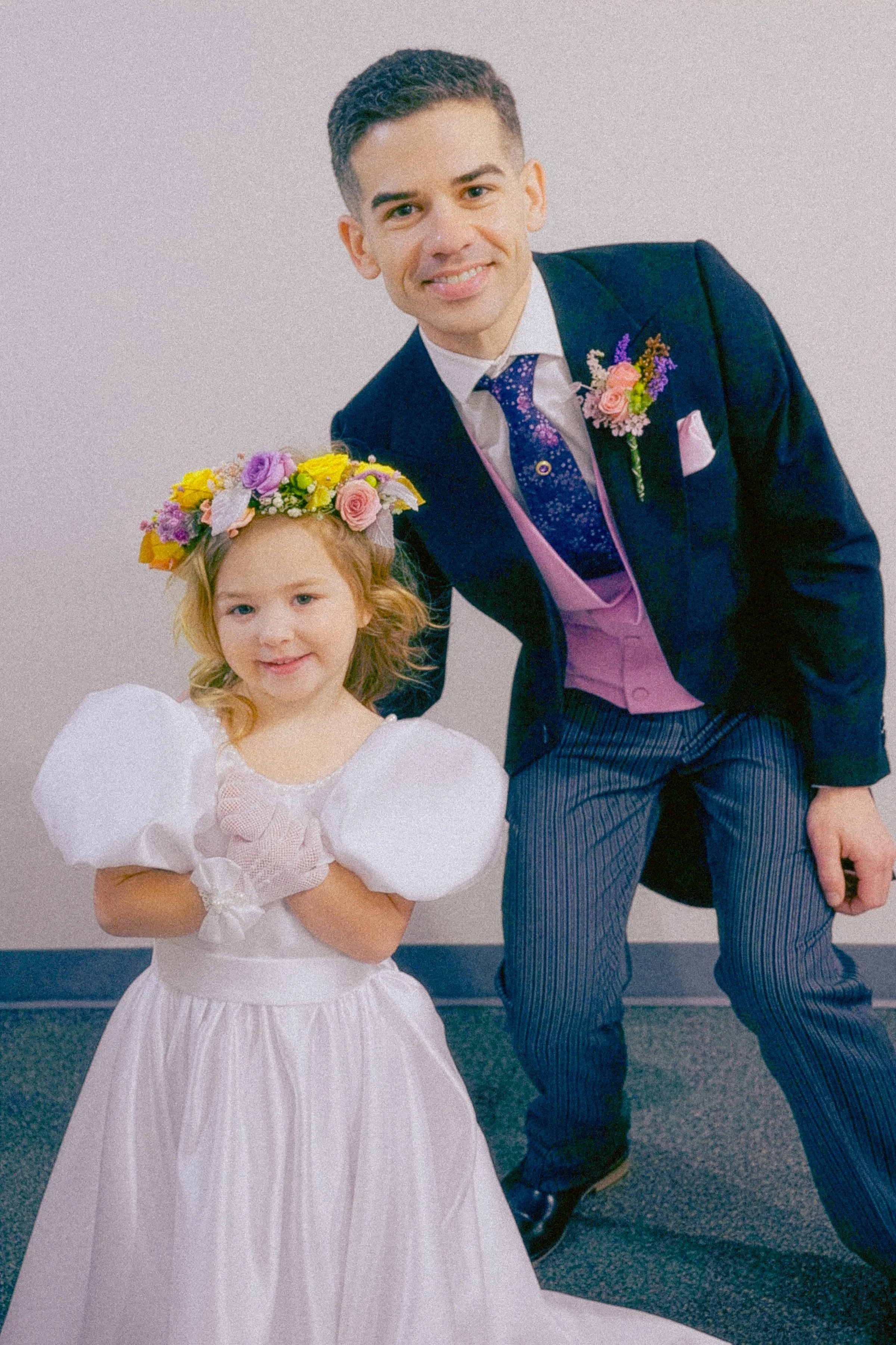 A young girl with curly hair wearing a white dress and a flower crown, standing in front of a man in a dark suit with a floral boutonniere at a formal event.