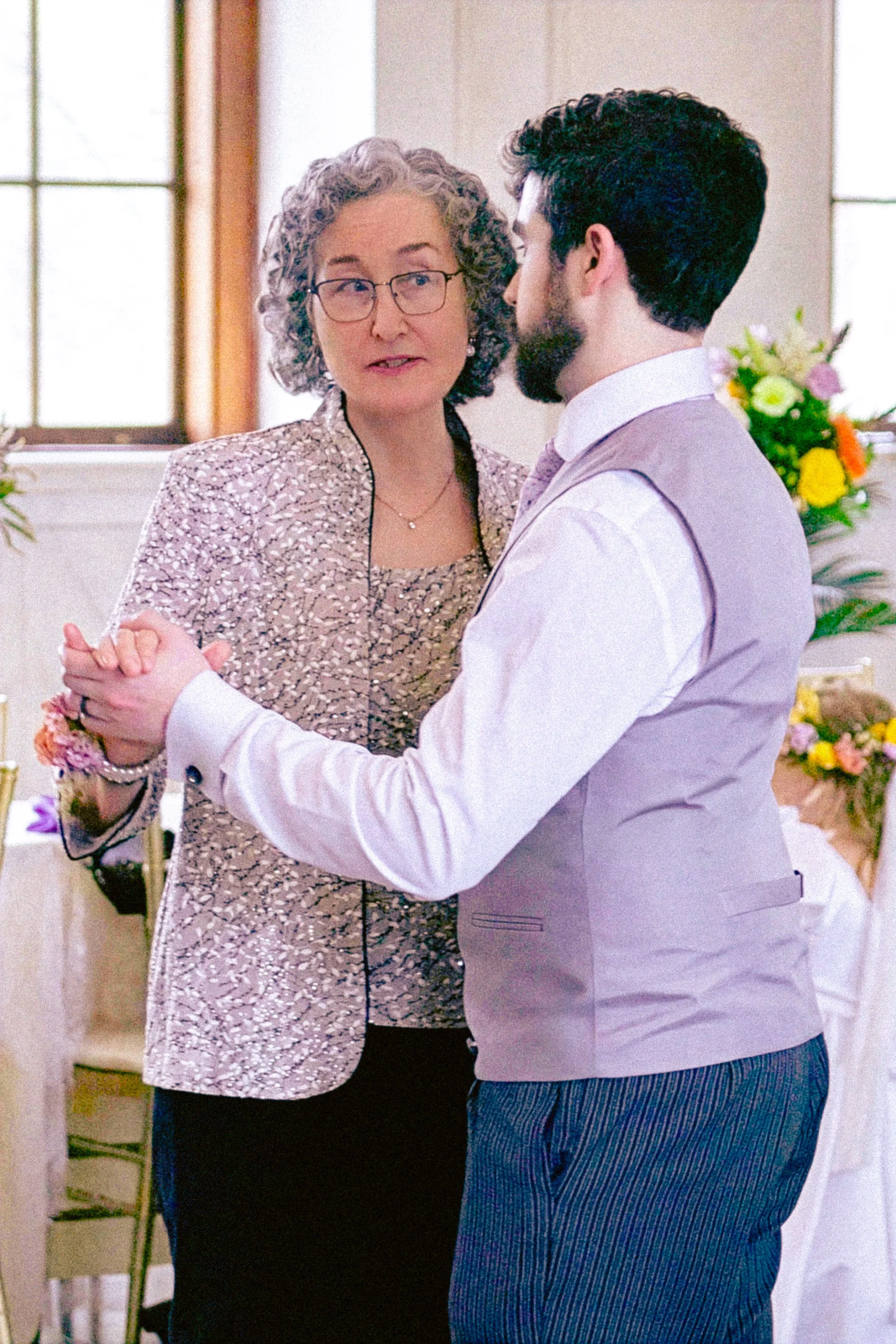 A woman with glasses and curly hair dancing with a man in a light gray vest and striped pants, holding hands at a formal event with floral decorations.