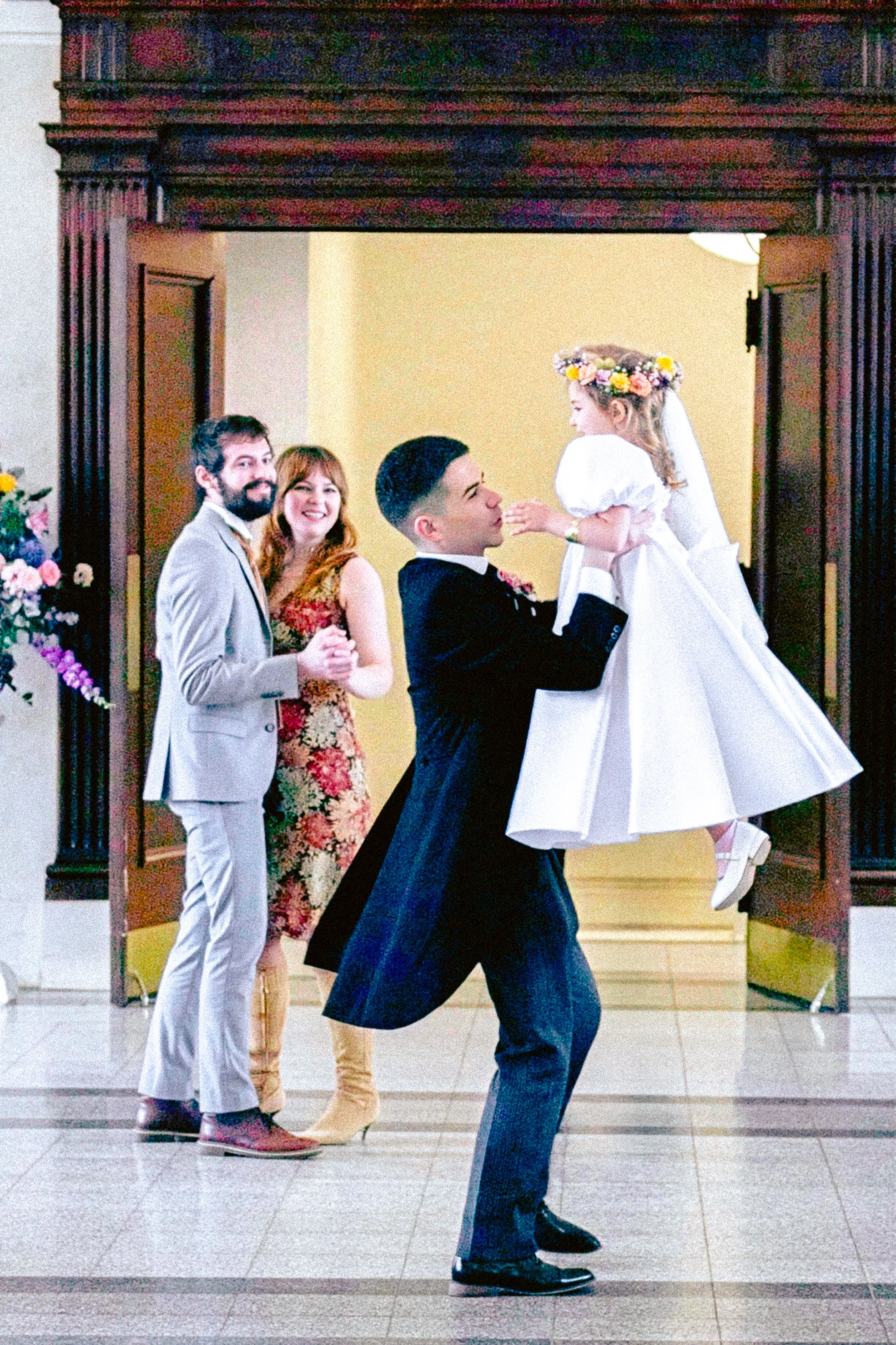 A group of four people, two men and two women, standing in a venue with wooden framing, engaged in a celebration. One woman is dressed in a wedding gown with a floral crown, and the man in front is holding her as if dancing. The others are smiling an
