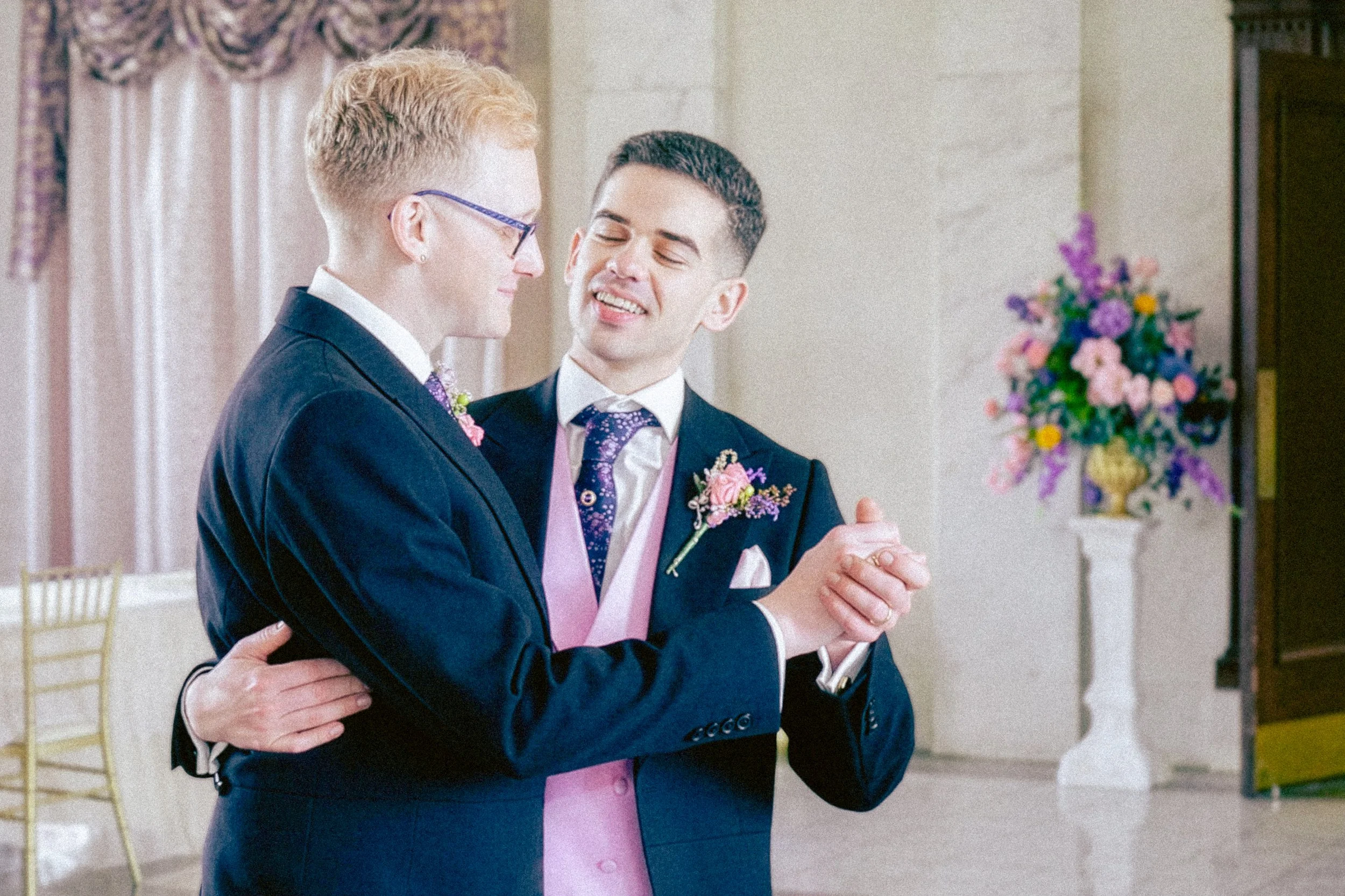 Two men at a wedding, dressed in dark suits with pink vests and purple ties, smiling and looking at each other, in a decorated indoor venue with floral arrangements in the background.