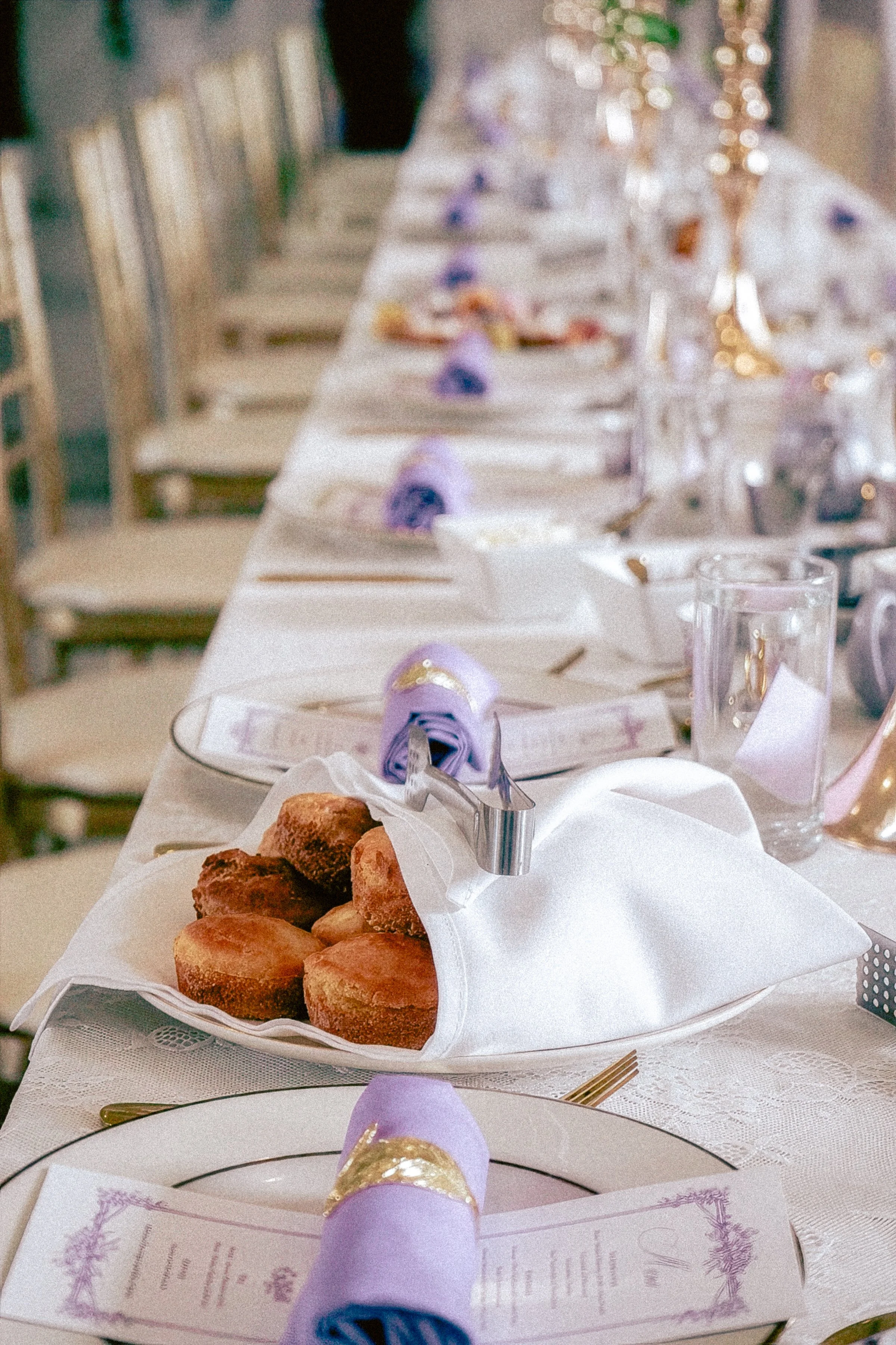 A banquet table set for an event with white tablecloths, decorated with purple napkins, gold accents, and wedding-style menus, with a plate of assorted cookies or petits fours in the foreground.