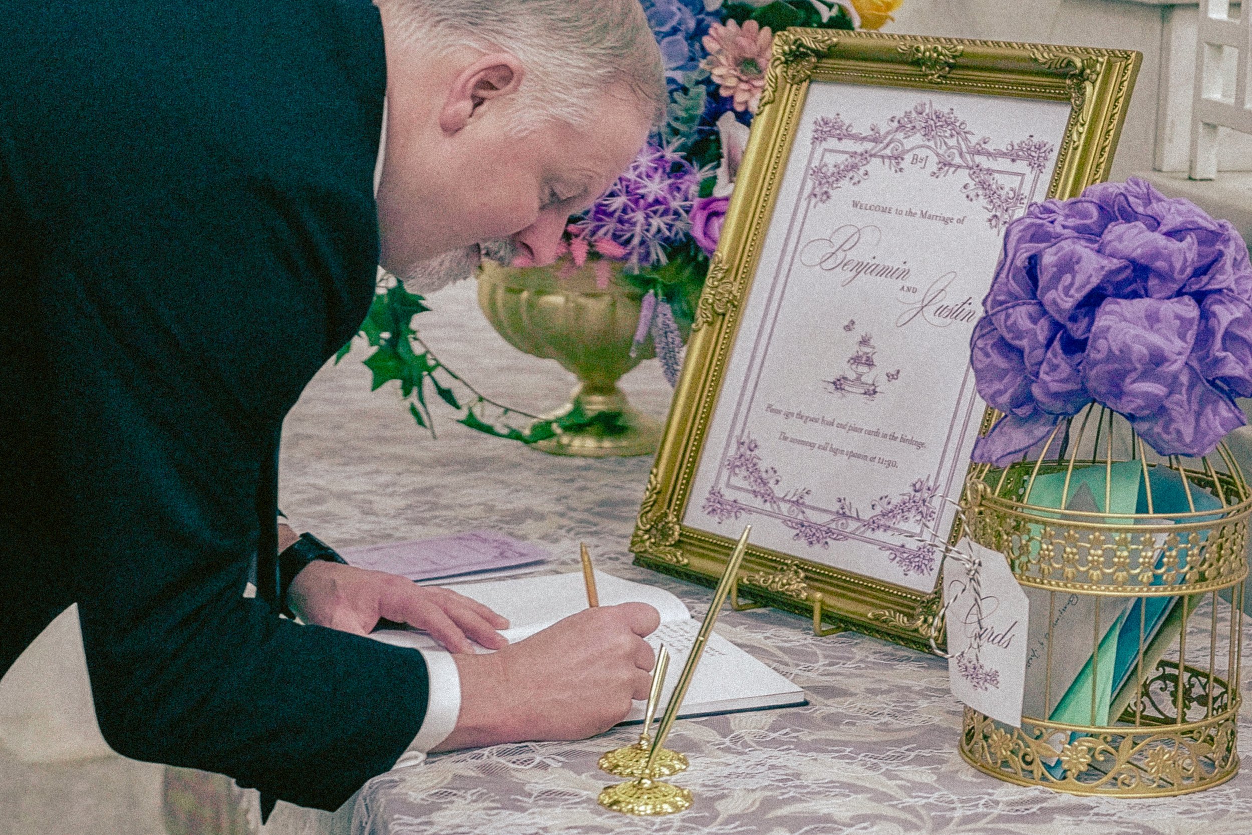 A man signing a marriage certificate at a wedding reception, with a decorative wedding sign and purple flower arrangements on the table.