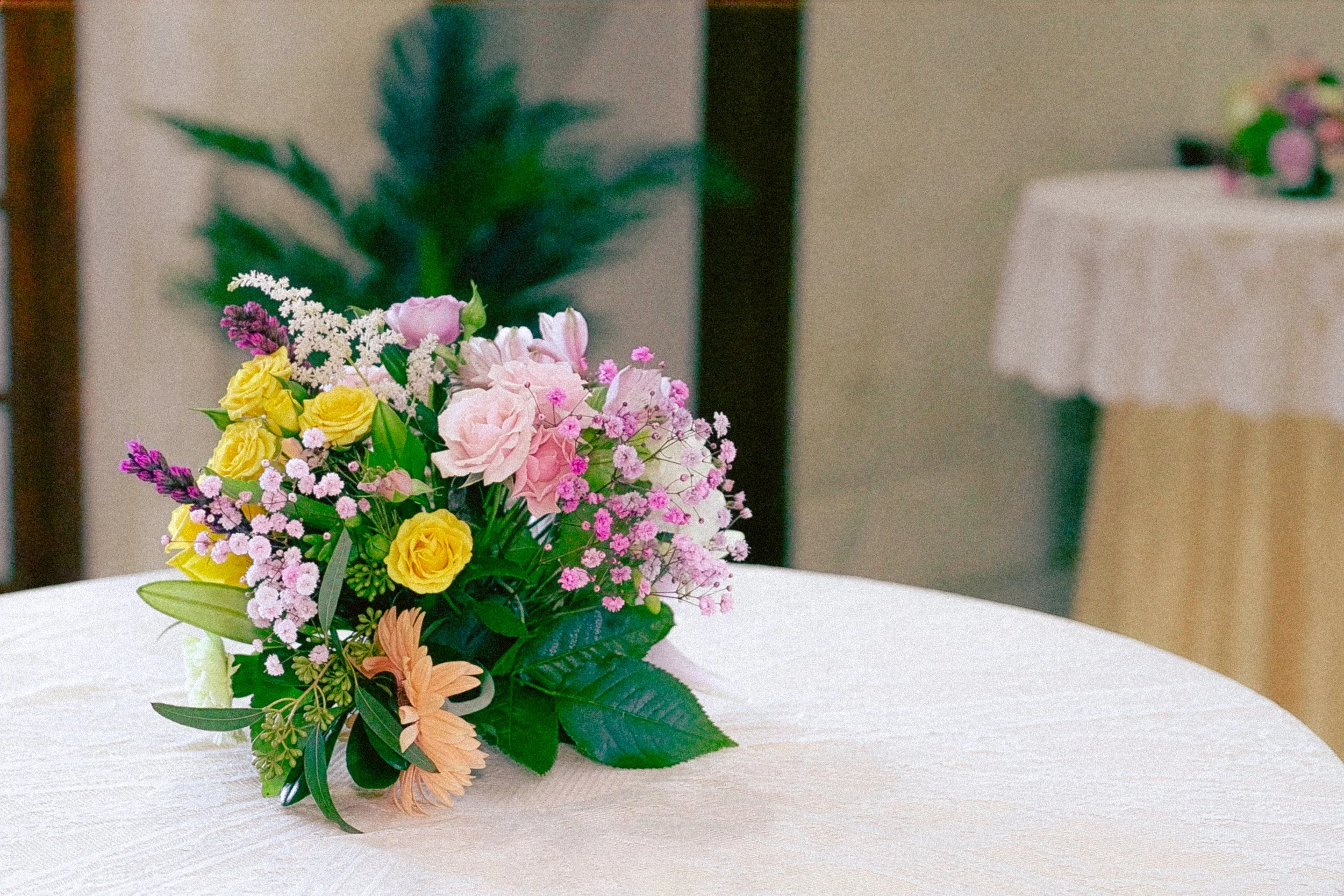 A bouquet of pink, yellow, white, and purple flowers arranged with green leaves, placed on a white table with a textured cloth. In the background, a large green plant and a blurred decorative item on a stand are visible.