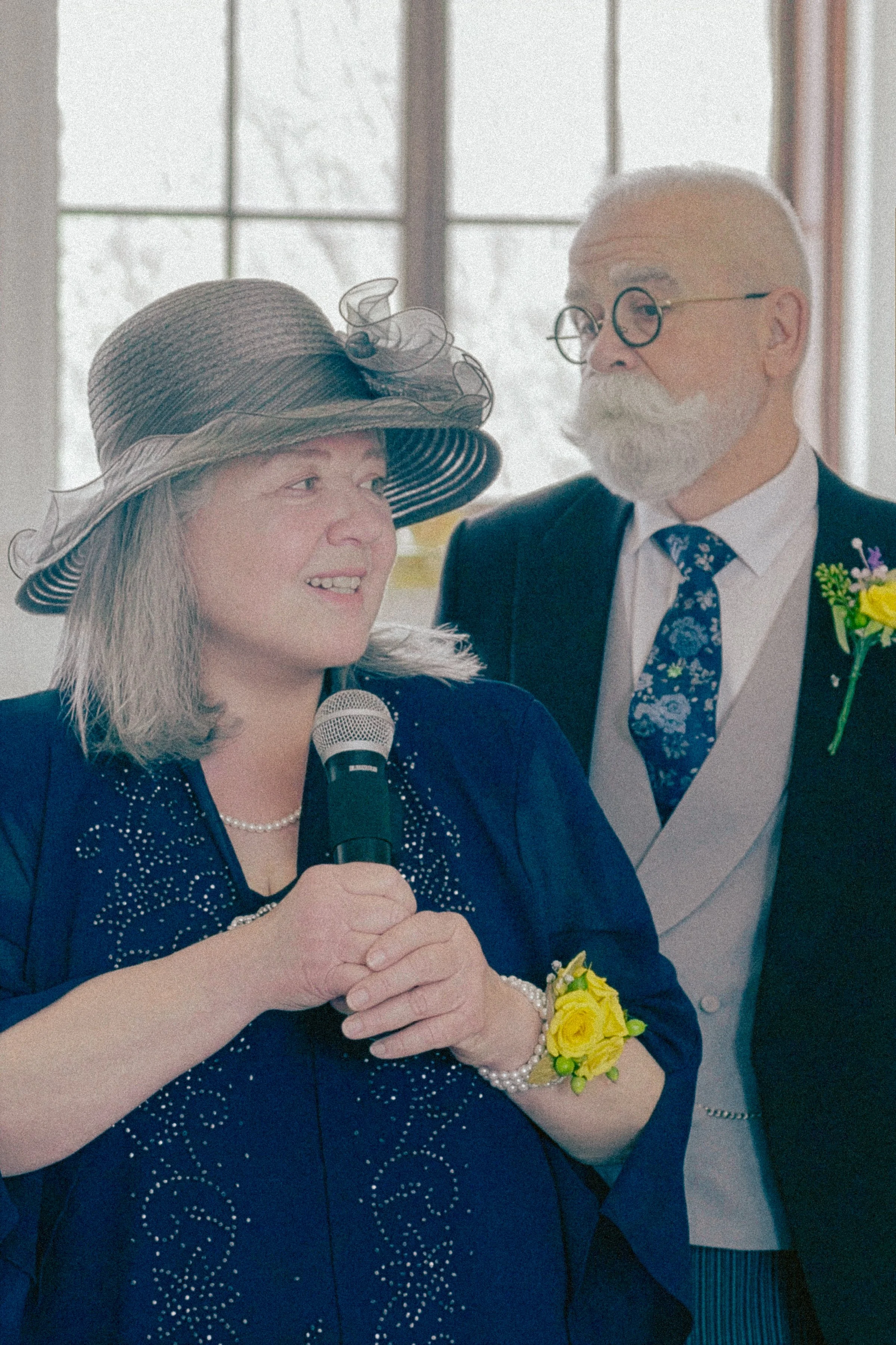 A woman wearing a large hat and a navy blue dress with pearl jewelry and a yellow flower bracelet, speaking into a microphone, with an older man in a suit and tie standing behind her indoors near a window.