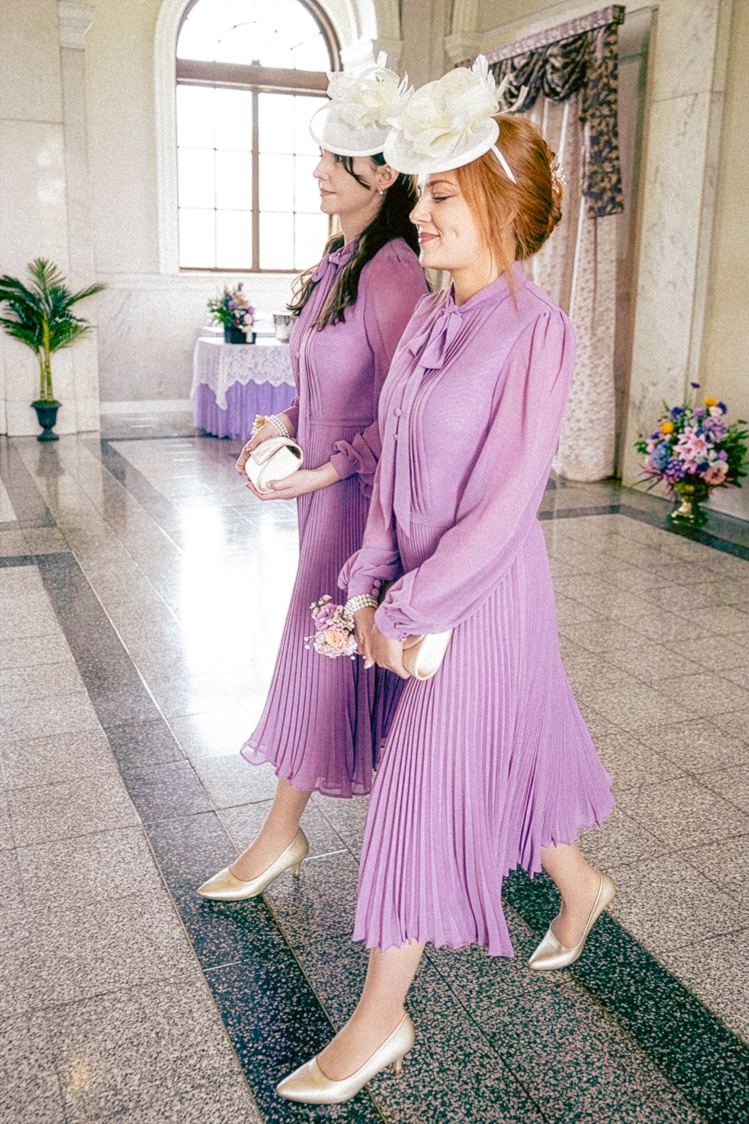 Two women in purple dresses and matching hats with large white flowers, walking side by side in an elegant indoor setting with floral arrangements and large windows.