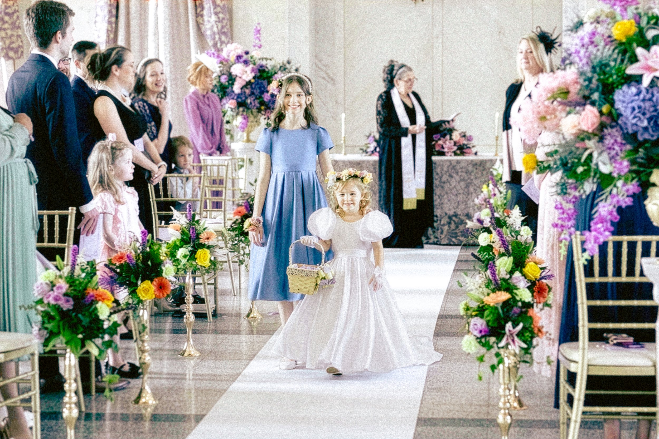 Two young girls walking down the aisle at a wedding ceremony, surrounded by colorful flowers and seated guests in a decorated indoor venue.