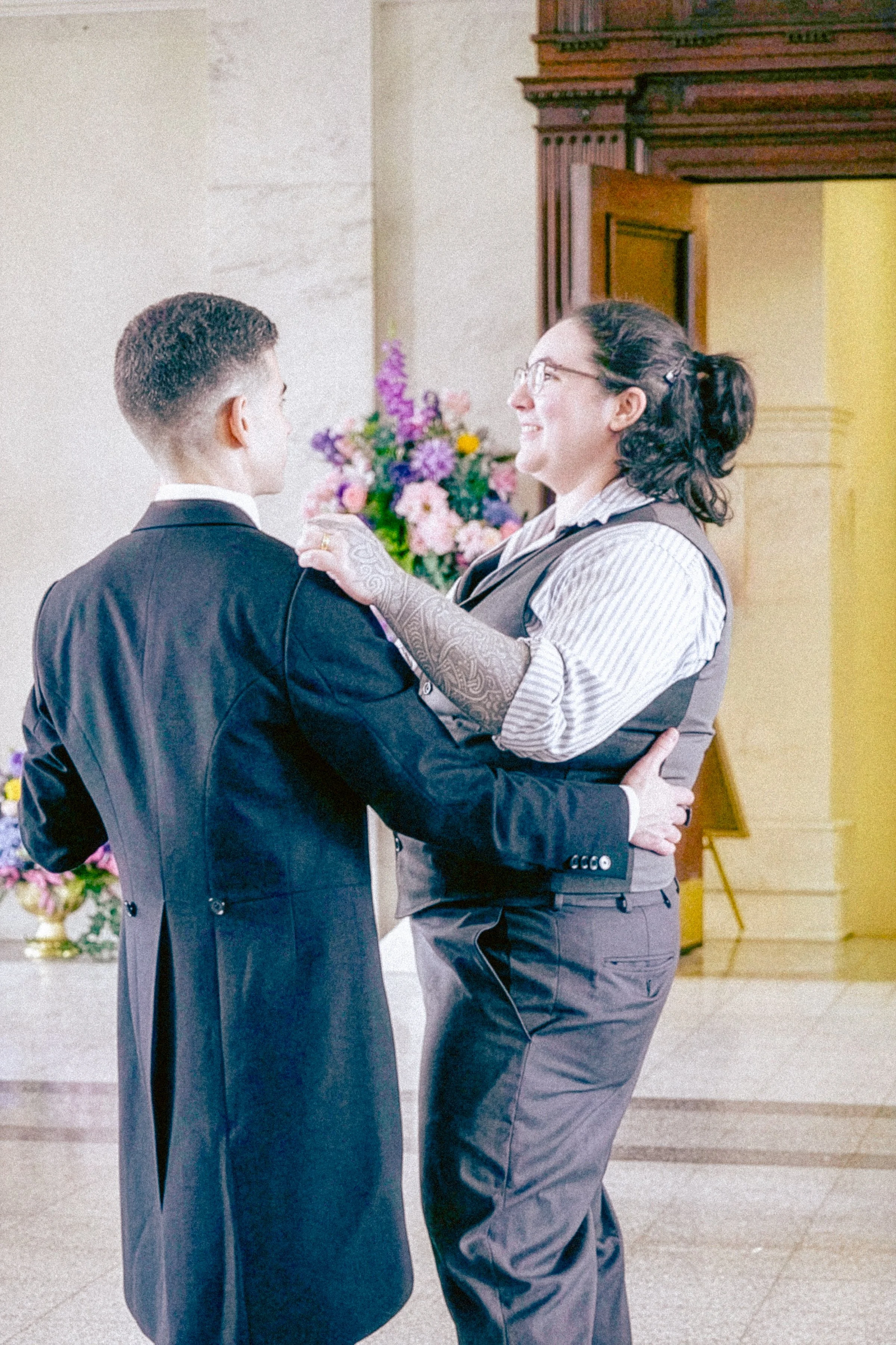 A woman with glasses and a tattoo on her arm is smiling and dancing with a man in a black suit during a celebration, with floral arrangements in the background.