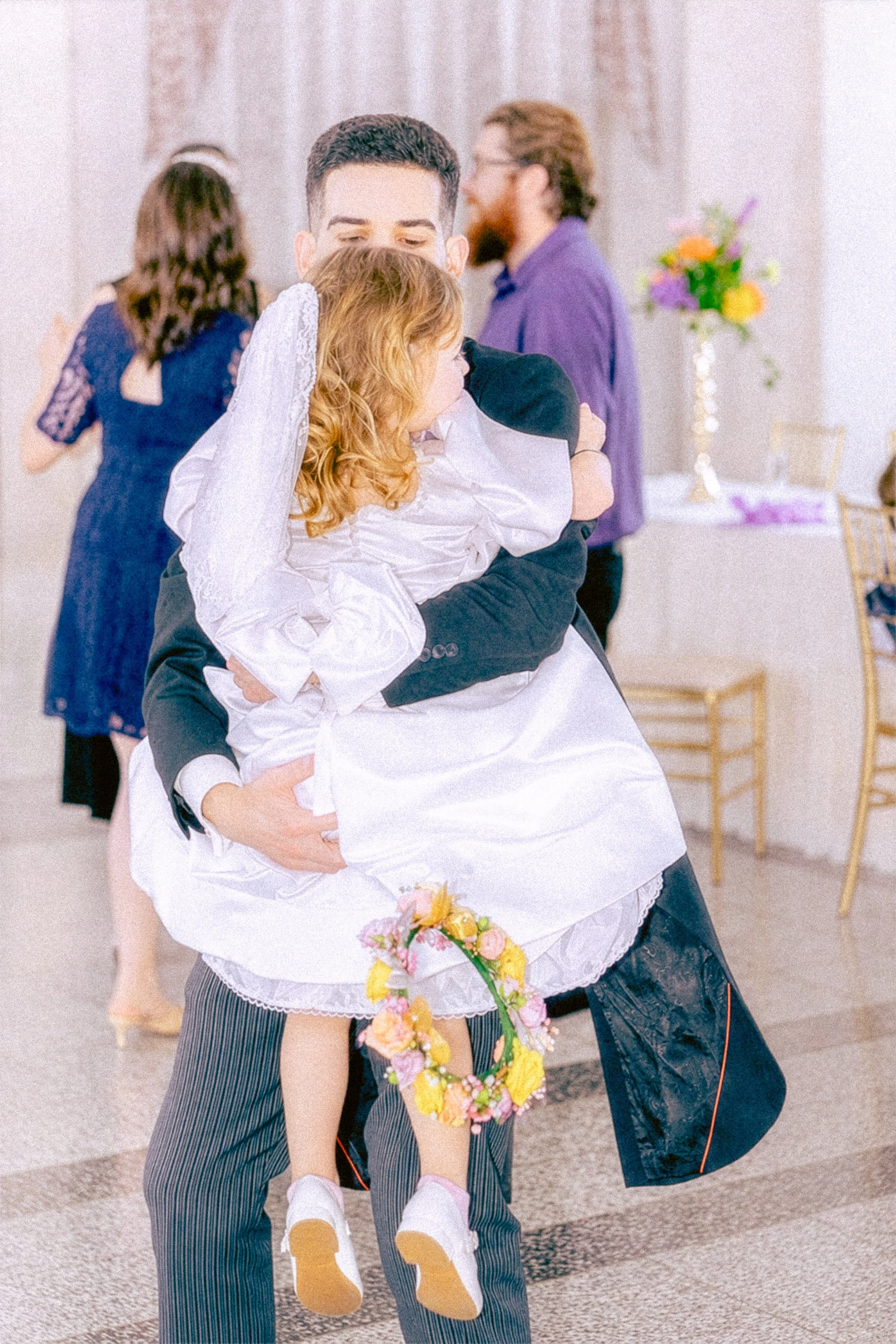 A couple dancing closely at a wedding reception, with a young girl holding a flower crown and wearing a white dress being lifted in the air. In the background, other guests are mingling, and a table with a colorful floral arrangement is visible.