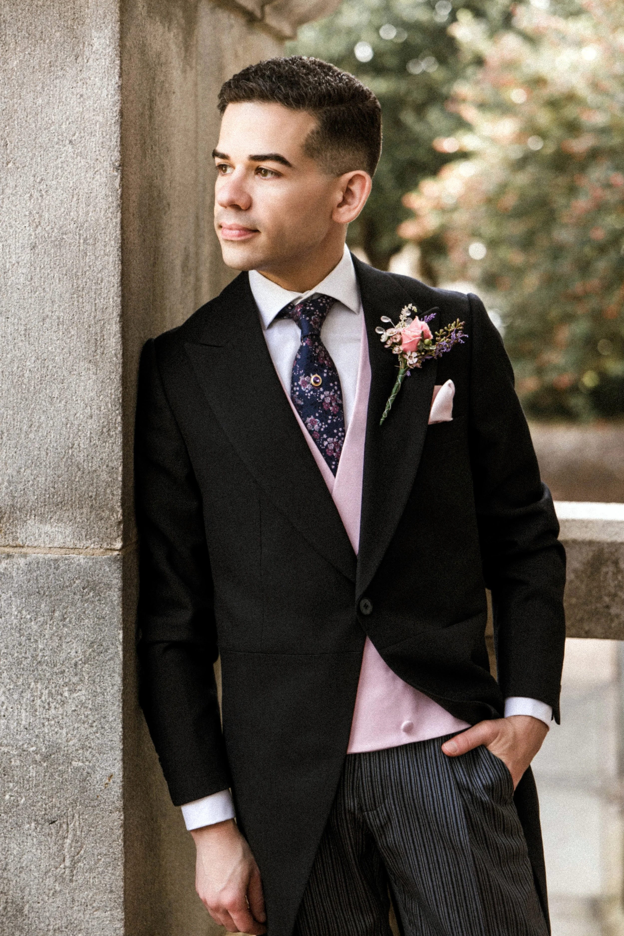 A man dressed in formal wedding attire, including a dark suit, white shirt, patterned tie, pink sash, and boutonniere, leans against a stone wall outdoors with trees in the background.