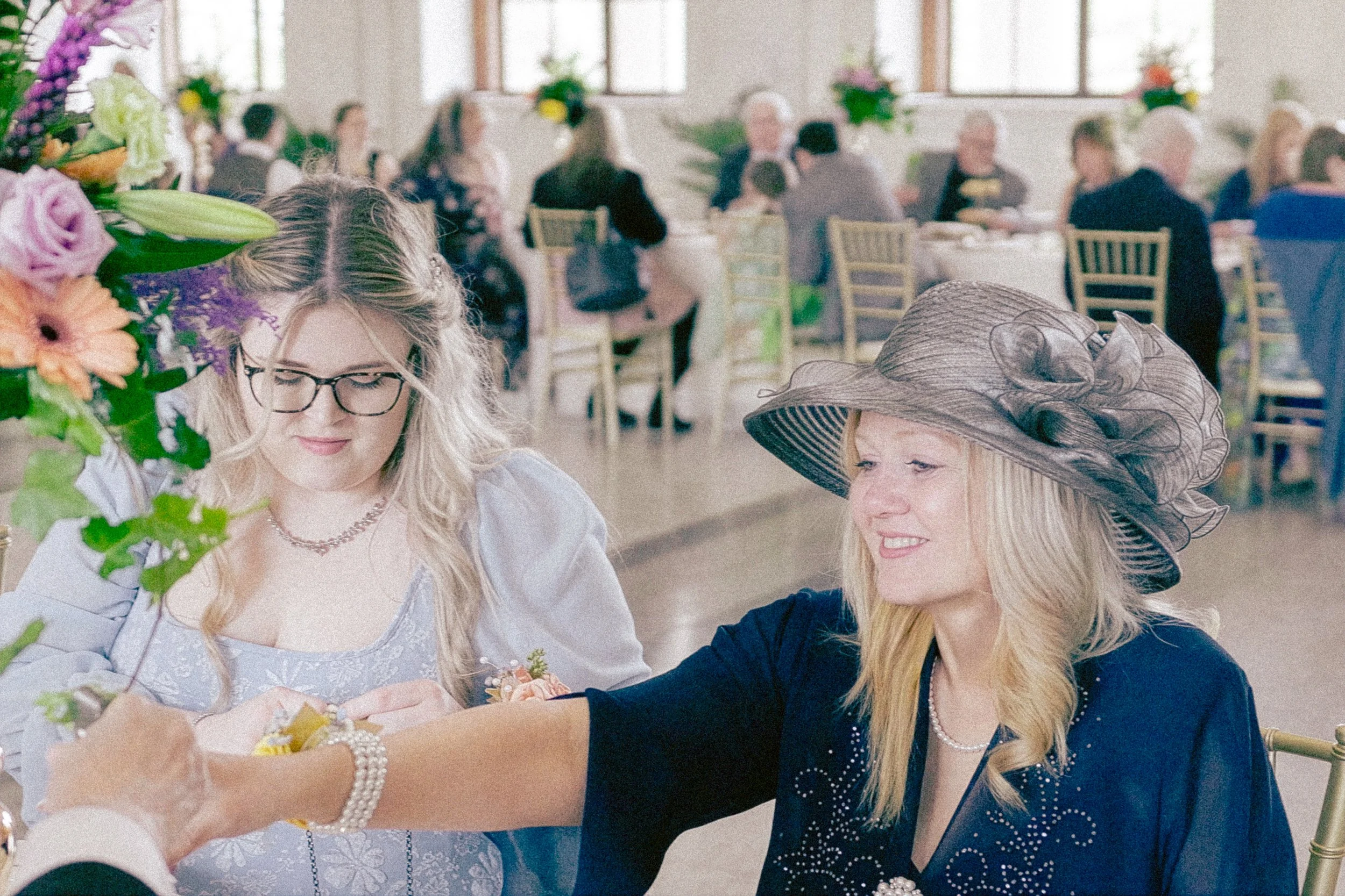 Two women, one with glasses and long blonde hair and the other wearing a large hat with decorative netting, are sitting at a table and exchanging gifts during a celebration or event. Behind them, a room filled with people seated at tables can be seen