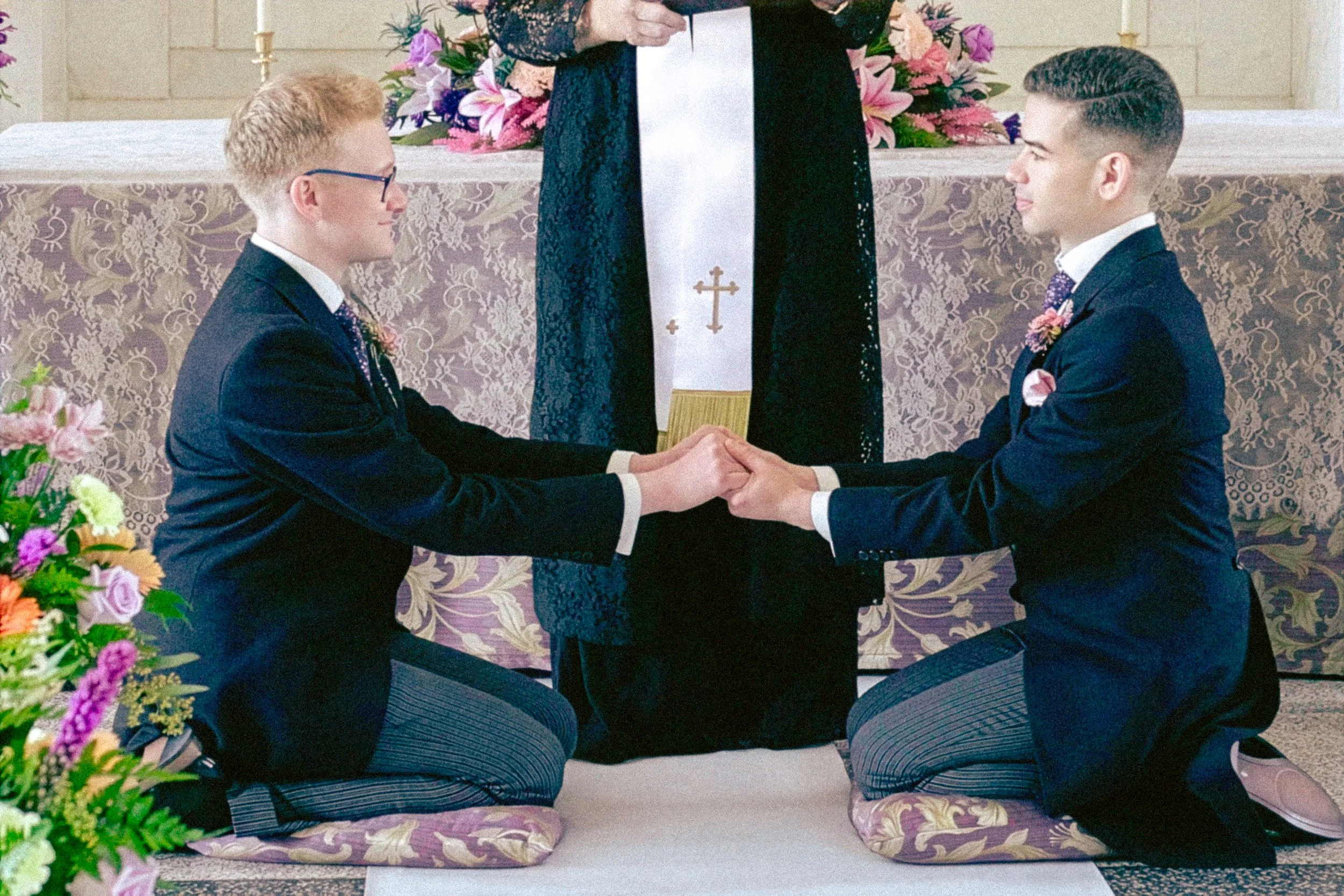 Two men kneeling and holding hands during a wedding ceremony, facing each other, with an officiant standing behind them holding a paper. The location is decorated with floral arrangements and a table with a cloth that has a cross symbol on it.
