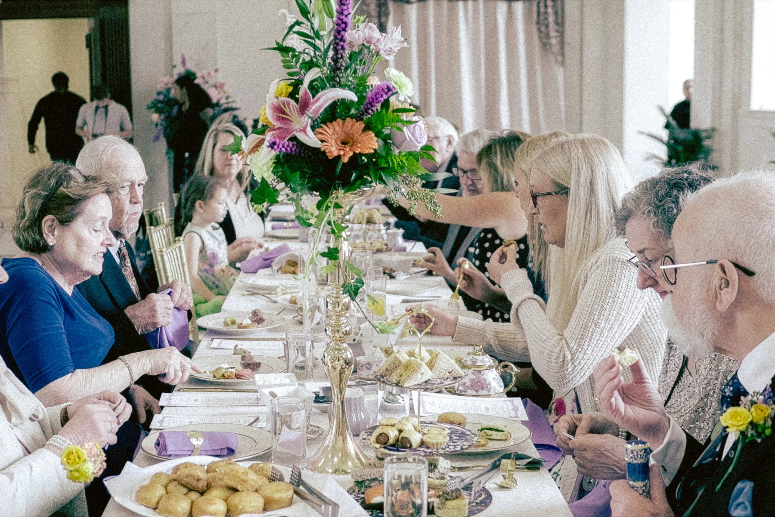 People seated at a long, elegant table during a formal gathering or celebration, with a large floral centerpiece featuring pink lilies, purple and white flowers, and greenery. The table is decorated with gold accents, and there are plates of assorted