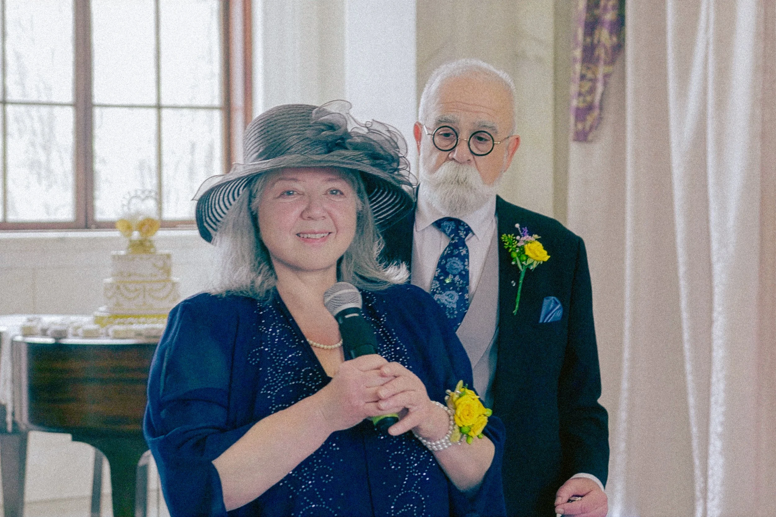A woman wearing a large sun hat and navy blue dress speaking into a microphone, with an older man in a suit and round glasses standing beside her, both smiling during a celebration or event in a decorated indoor setting.