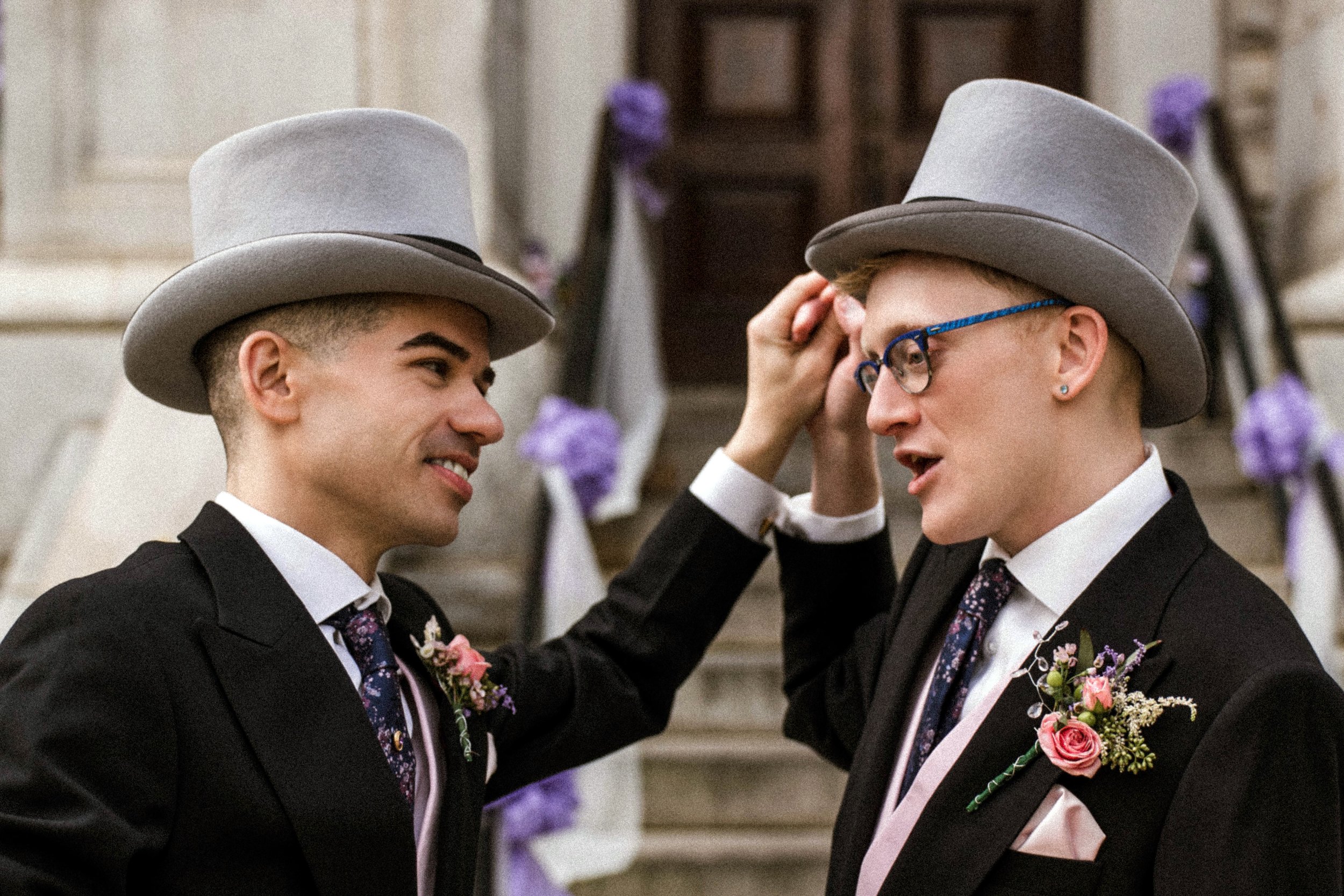 Two men in formal suits and gray bowler hats engaging in a conversation at a wedding or formal event. One man is adjusting the other’s hat.