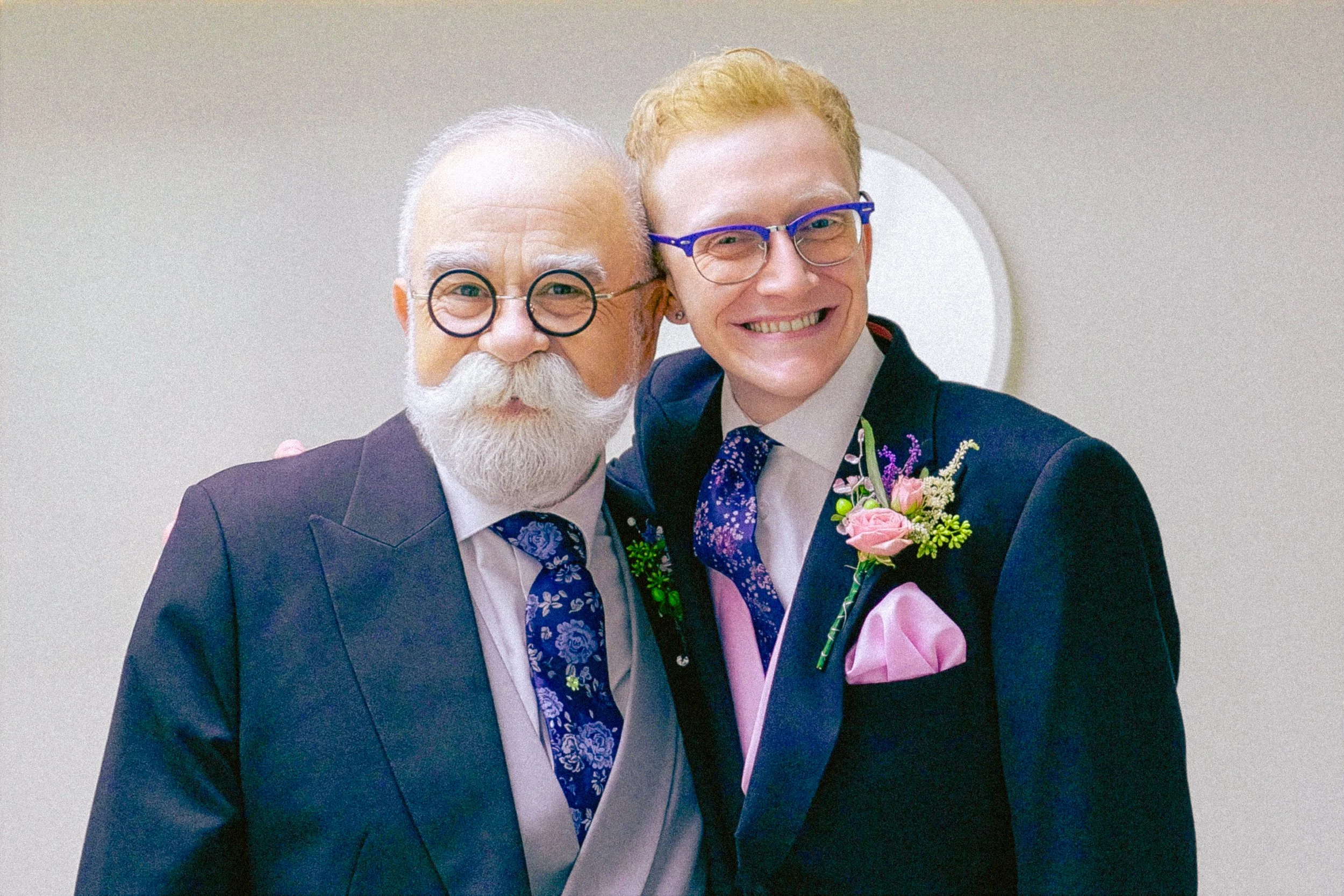 Two men dressed in formal suits smiling for a photo, with boutonnières and colorful ties, standing against a plain wall.