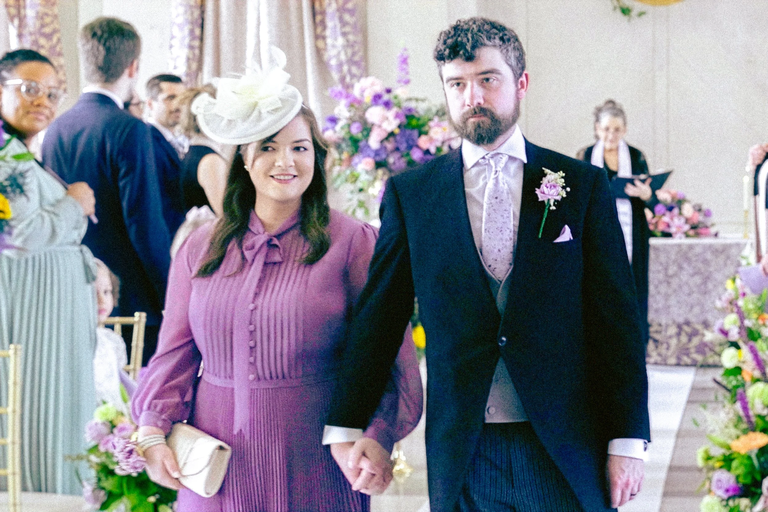 A couple dressed in formal attire holding hands at a wedding ceremony. The woman is wearing a pink dress with a matching hat and the man is in a dark suit with a light-colored tie and boutonniere. They are surrounded by guests and floral decorations.