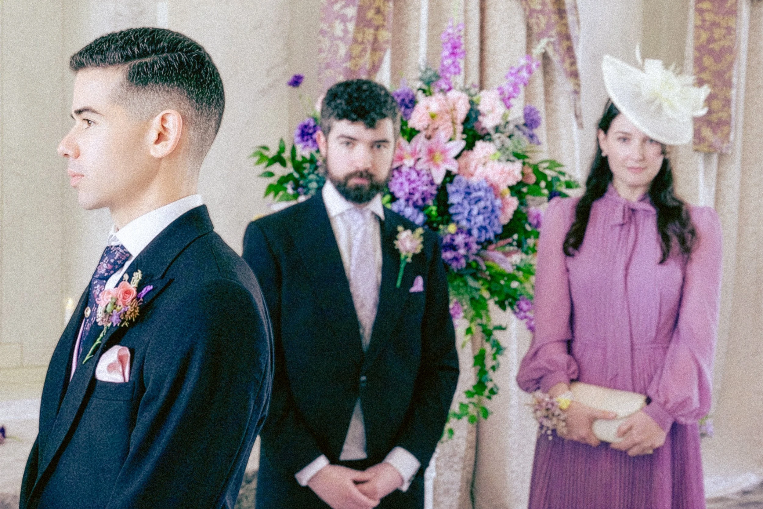 Group of three people, two men and a woman, dressed in formal attire, standing in front of a floral backdrop.