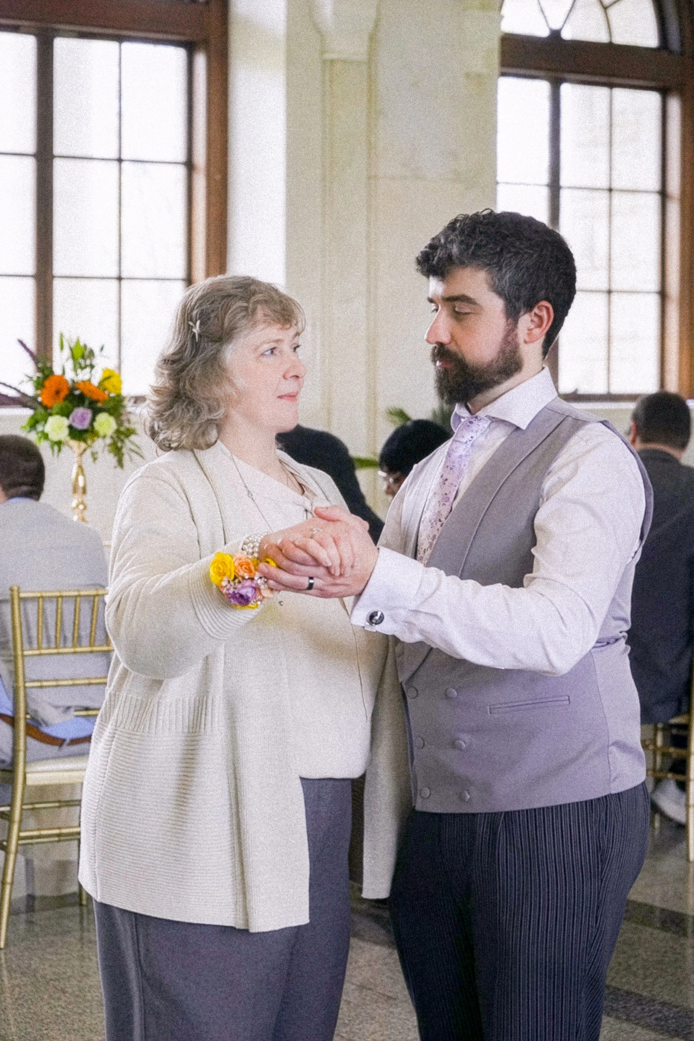 A middle-aged woman and a young man dancing together at a formal event, holding hands and looking into each other's eyes, with other guests and large windows in the background.