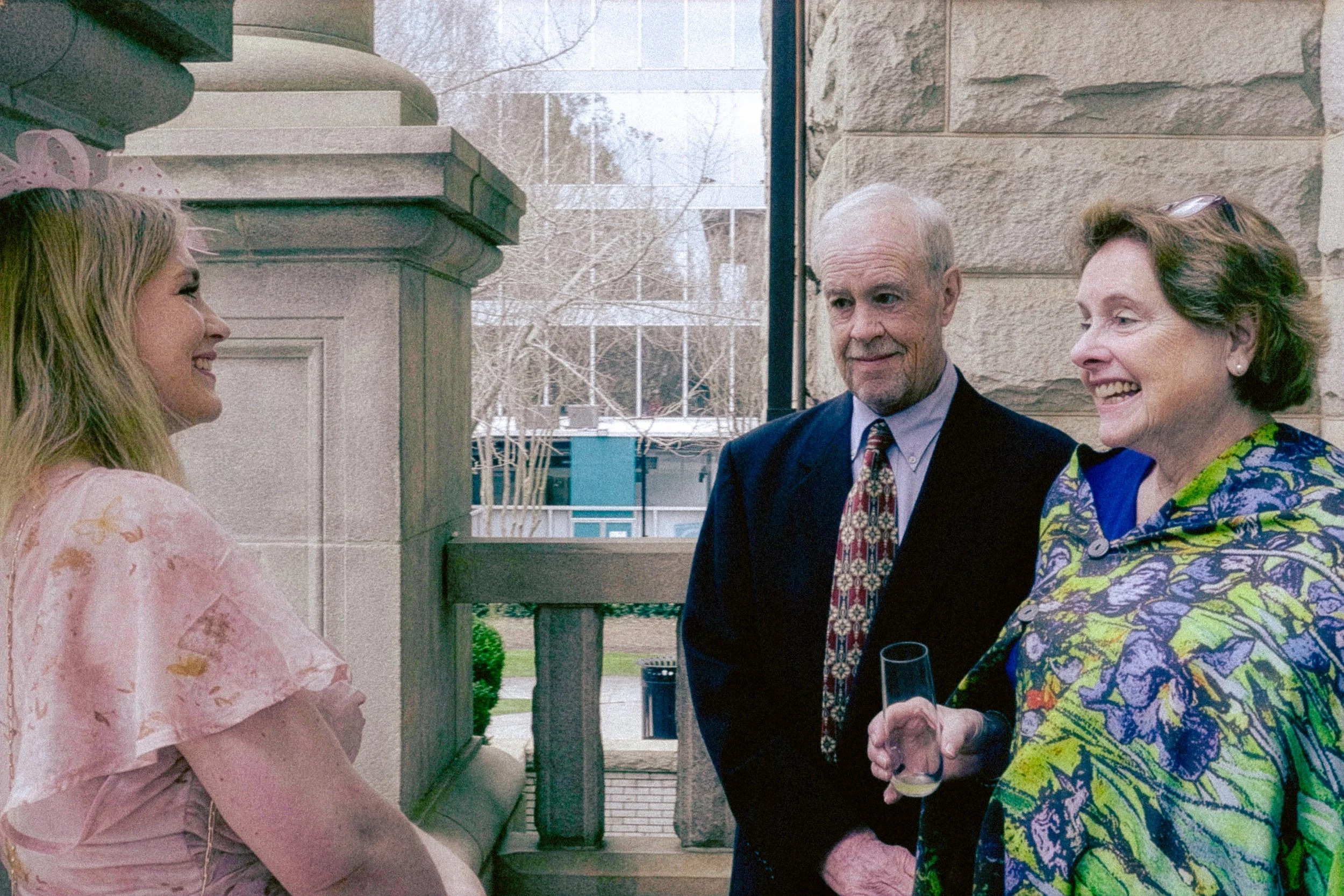 Three elderly people and a young woman interacting outdoors on a porch or patio of a stone building during daytime. The elderly man in the center, wearing a dark suit, tie, and light shirt, is smiling and holding a glass of wine. The elderly woman on