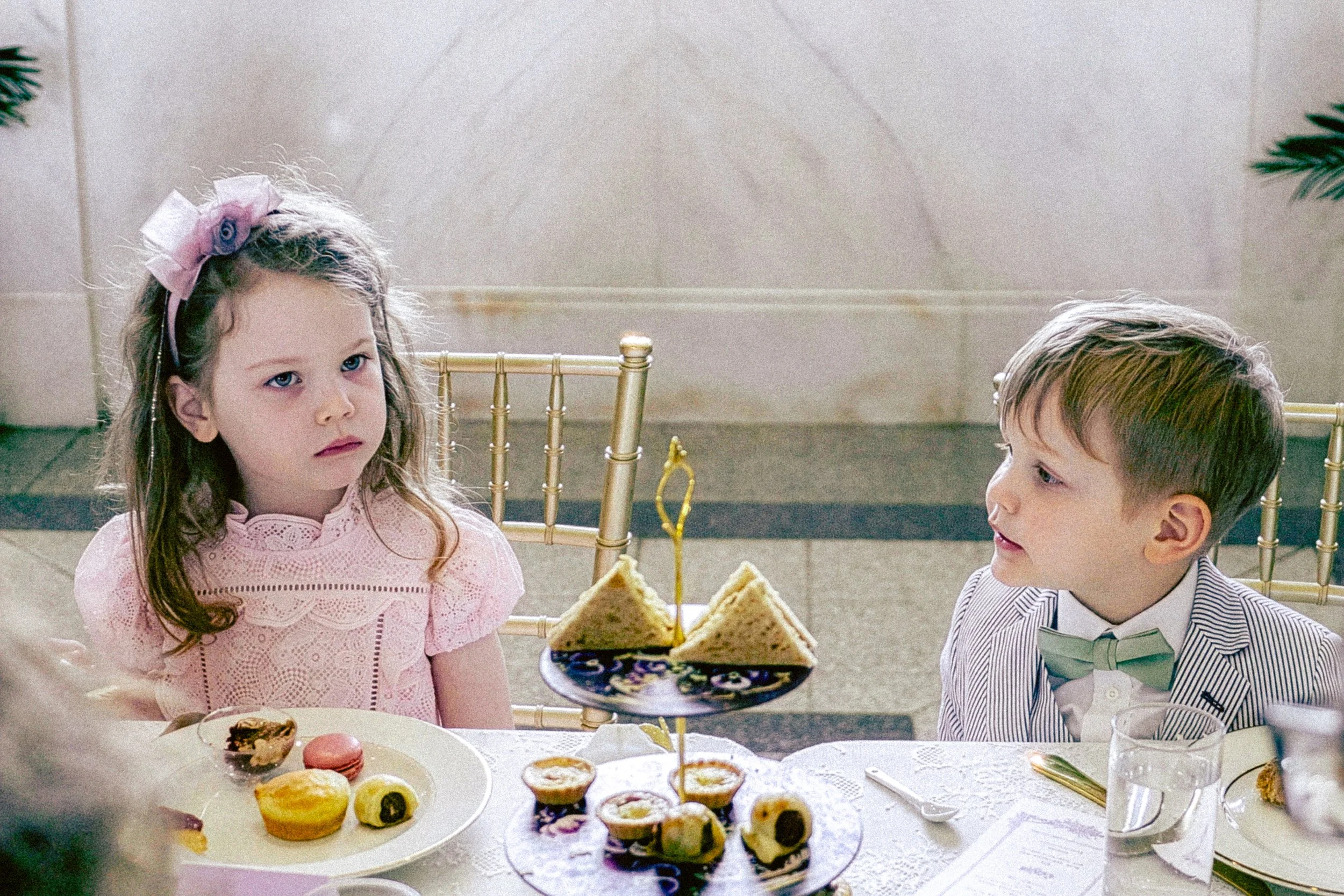 Young girl with wavy hair and a pink bow sitting at a table with a plate of assorted desserts. Another young boy with short hair and a striped shirt is sitting next to her. There is a multi-tiered tray of cupcakes and party food on the table, and the