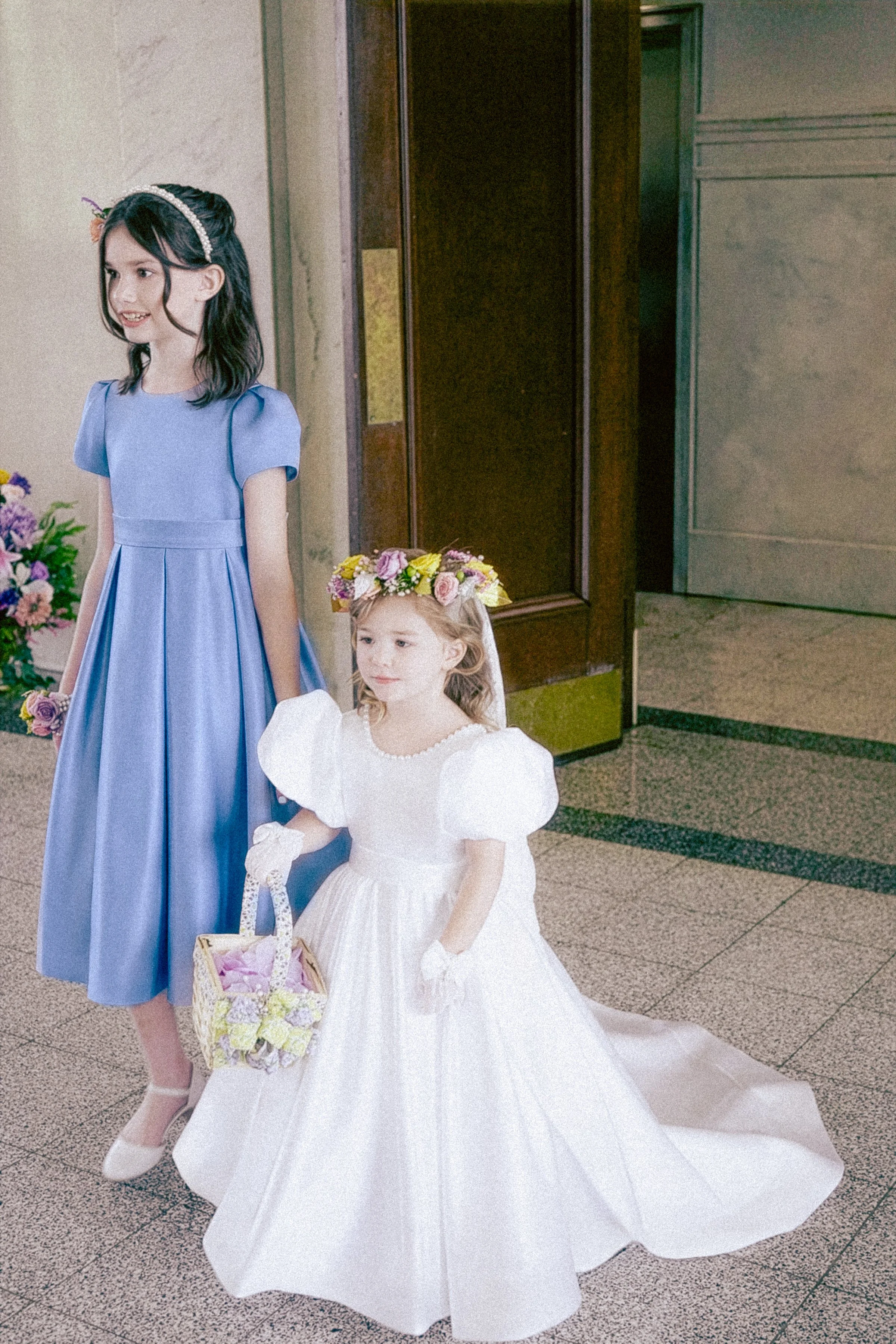 A young girl in a white dress and gloves holding a basket, standing next to a woman in a blue dress with a flower crown and pearl headband, in an indoor setting with floral arrangements nearby.