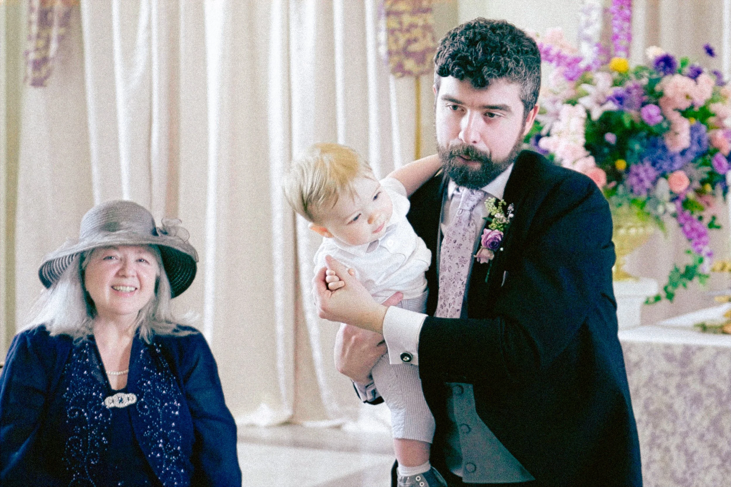 A man in a dark suit holding a young boy with blond hair at a celebration, next to a woman wearing a large hat and dark velvet dress, with colorful flowers in the background.