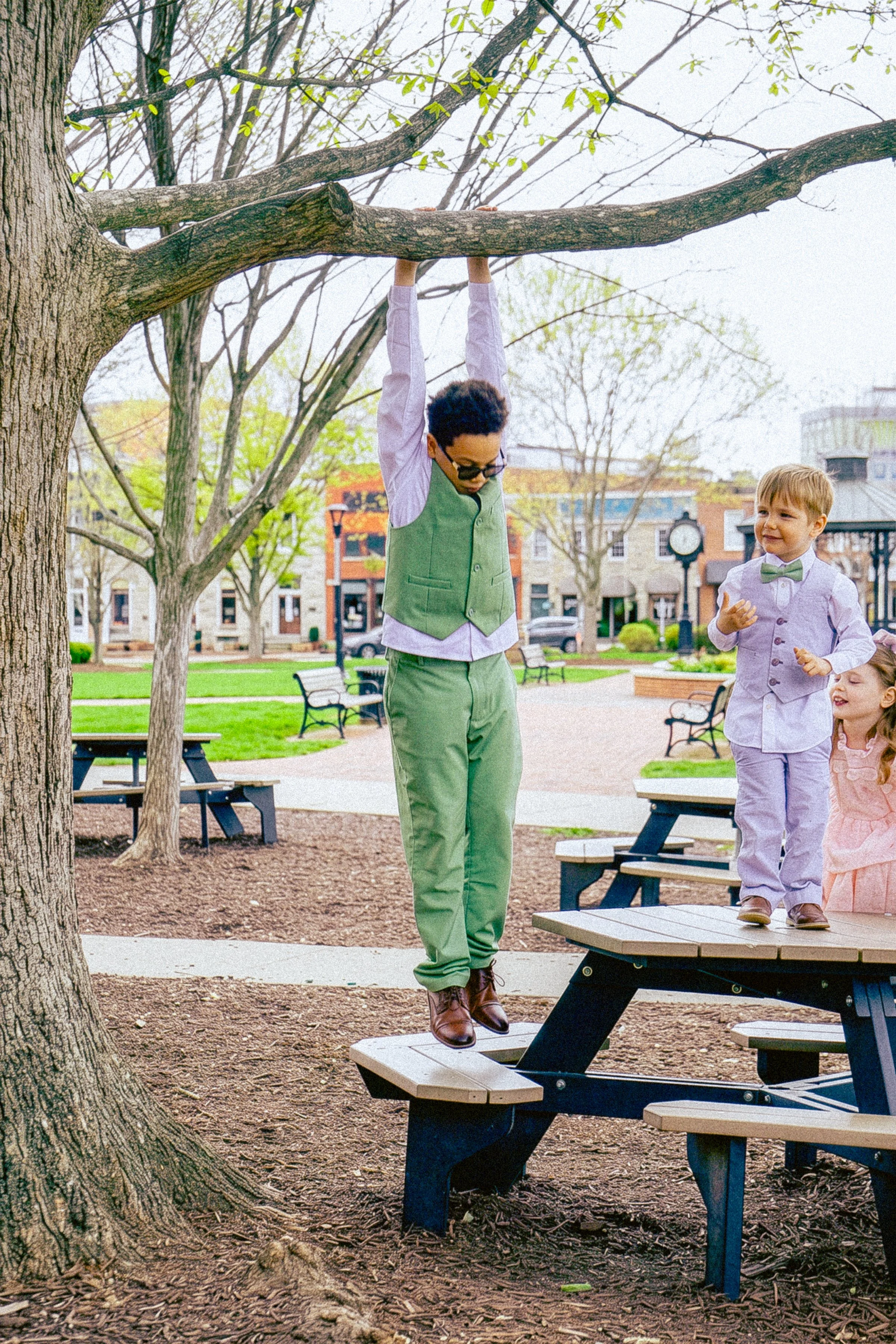 Children and adult walking on picnic tables in park with trees and buildings in background.