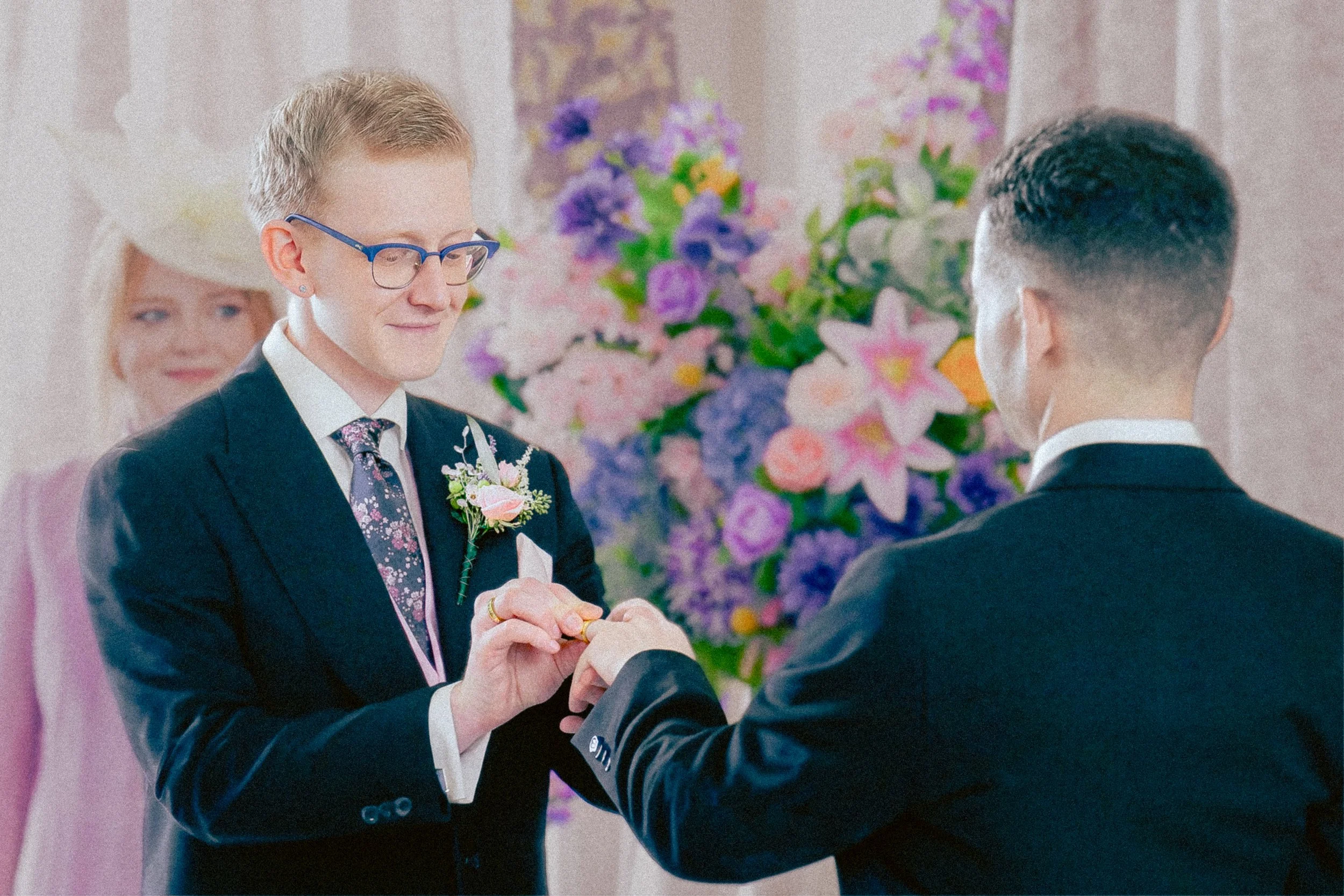 A man with glasses and a floral tie exchanges rings with another man during a wedding ceremony, with a woman smiling in the background and colorful floral arrangements in the setting.