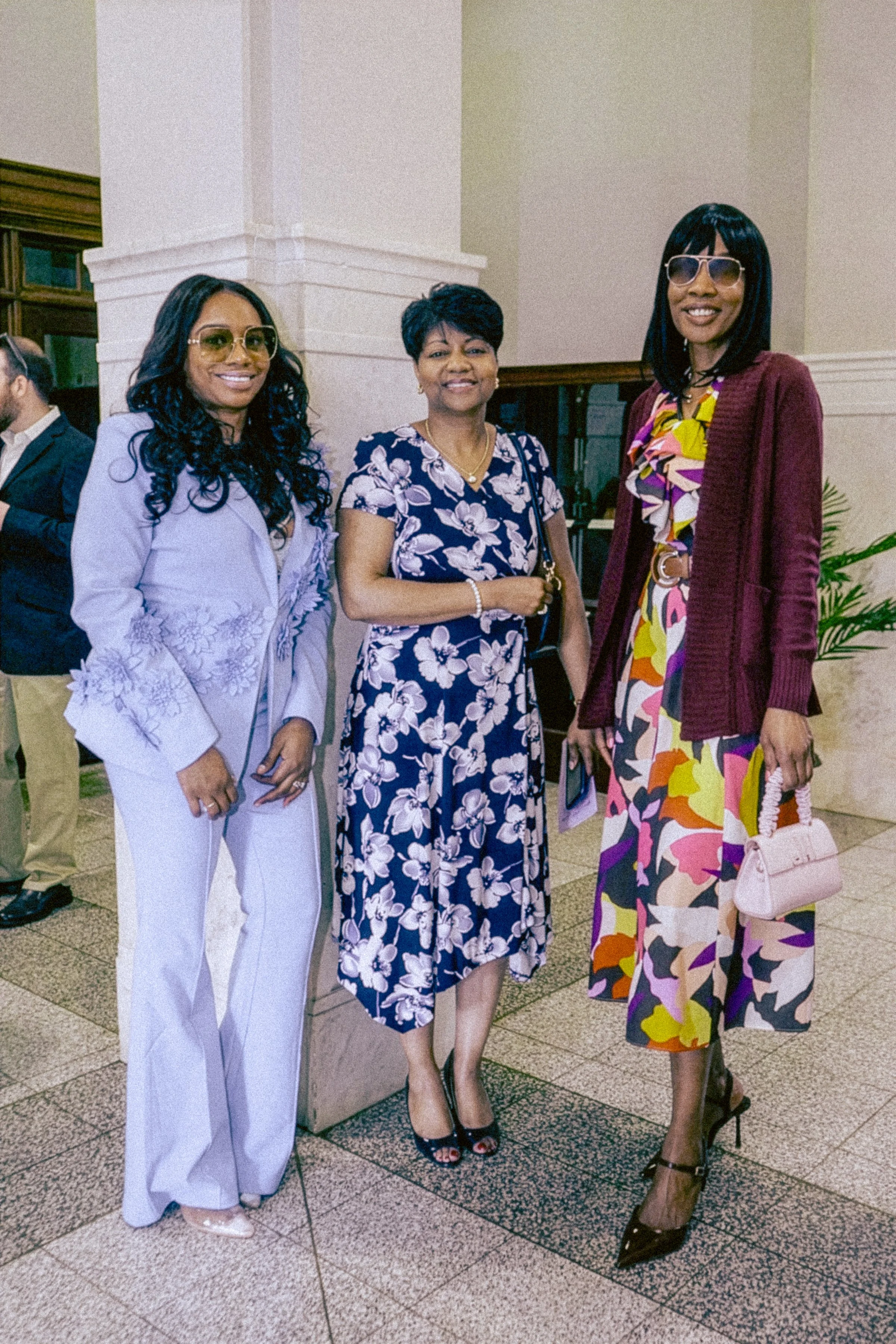 Three women standing together indoors, all smiling. The woman on the left wears a light purple suit with lace details and yellow-tinted glasses. The woman in the middle is dressed in a floral navy blue dress, holding a small clutch, with short dark h