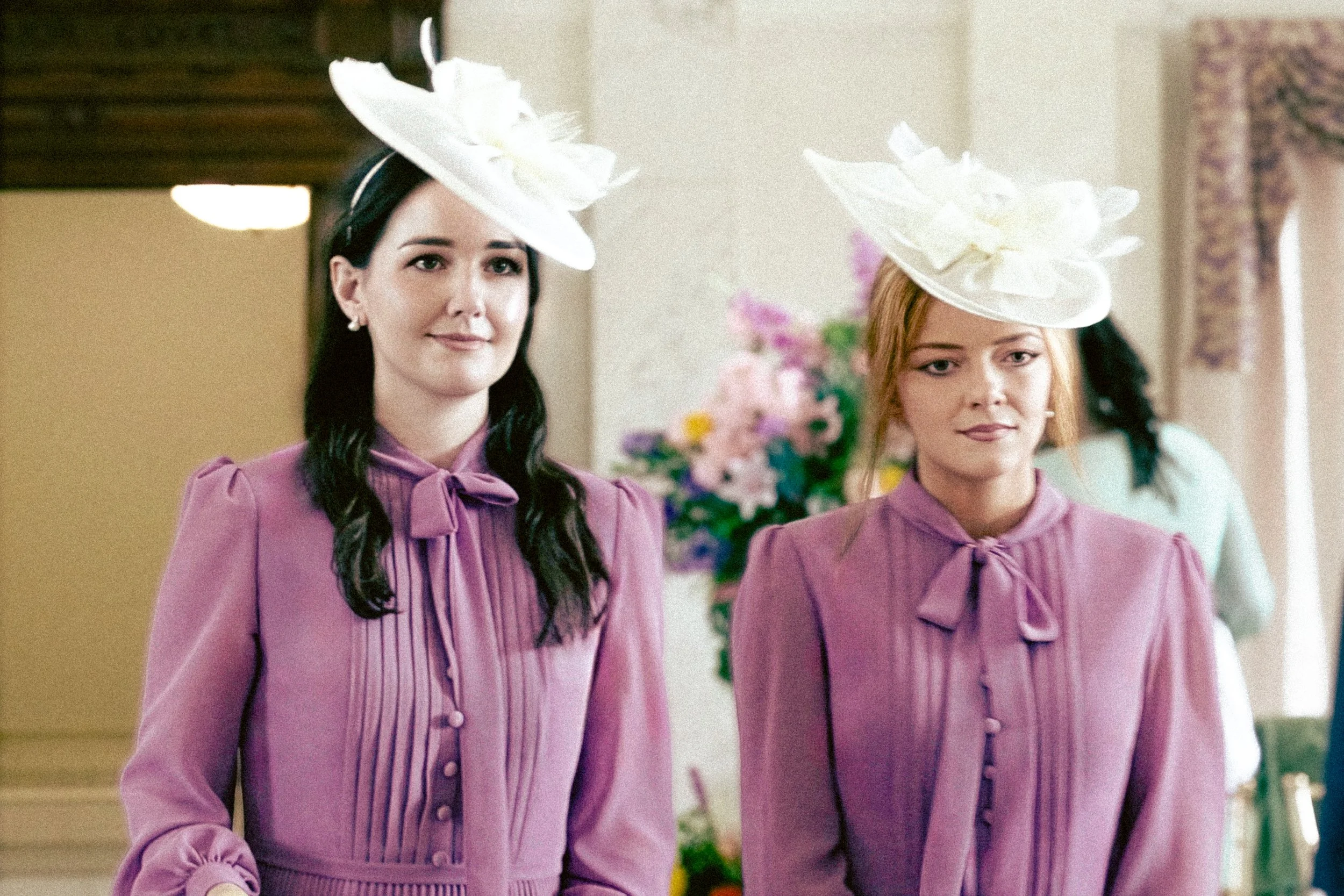 Two women wearing matching purple dresses with pleated fronts and decorative buttons, along with large white hats adorned with fabric flowers, standing indoors with floral decorations in the background.