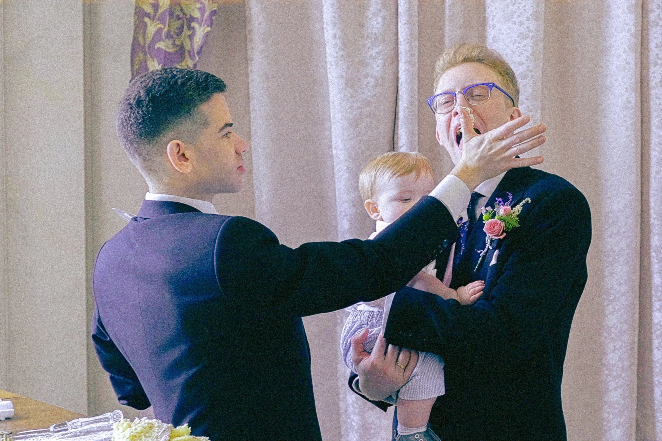 Two men in formal suits, one with glasses, are participating in a wedding ceremony. One man is holding a young girl, who has red hair and is wearing a light-colored dress. The man with glasses is yawning or stretching, with his hand over his face, wh