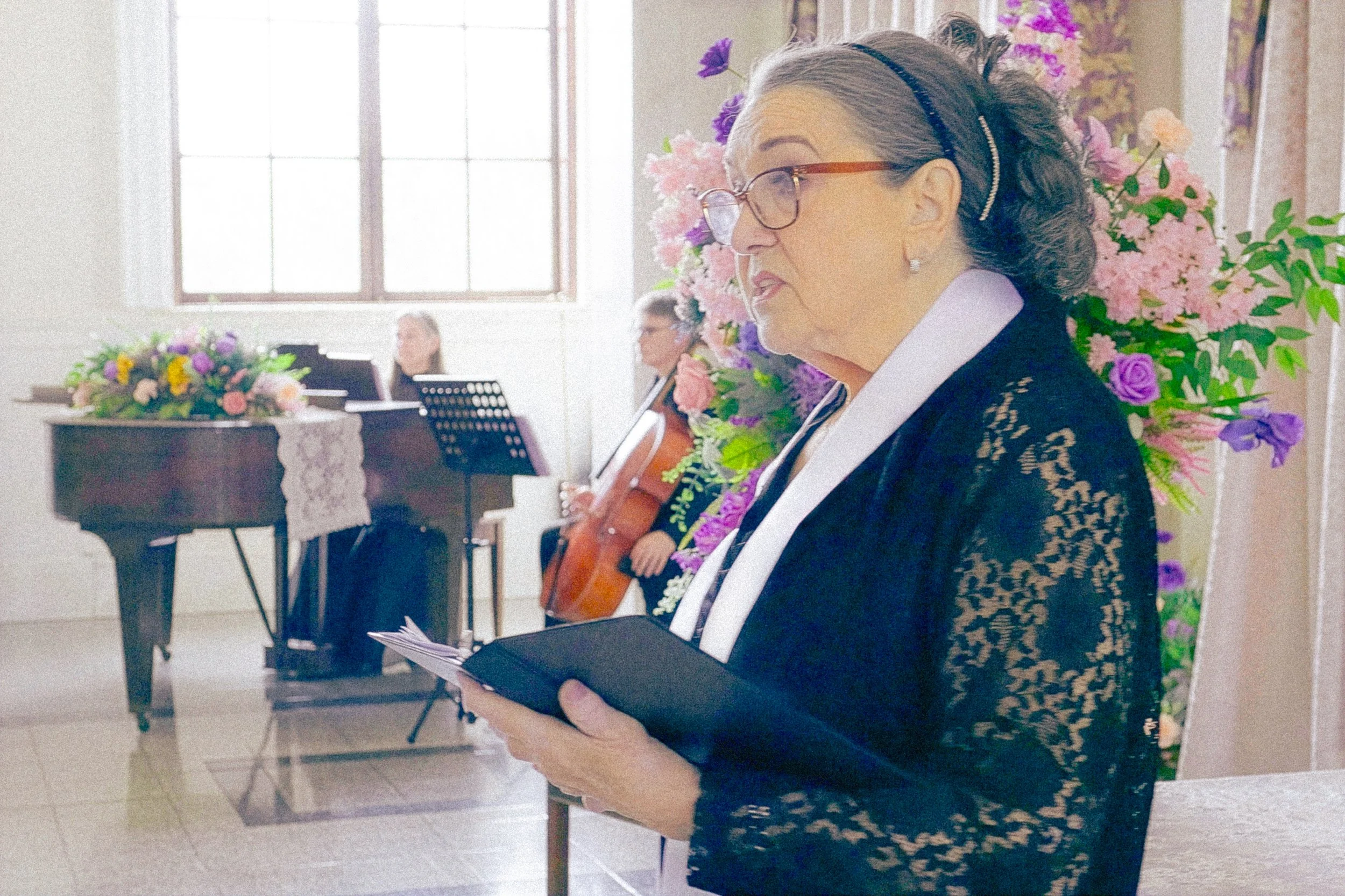 An elderly woman with glasses and gray hair styled in curls, wearing a black lace dress, is holding a booklet or program. She is in a room decorated with numerous pink and purple flowers, and there is a piano with a floral arrangement in the backgrou
