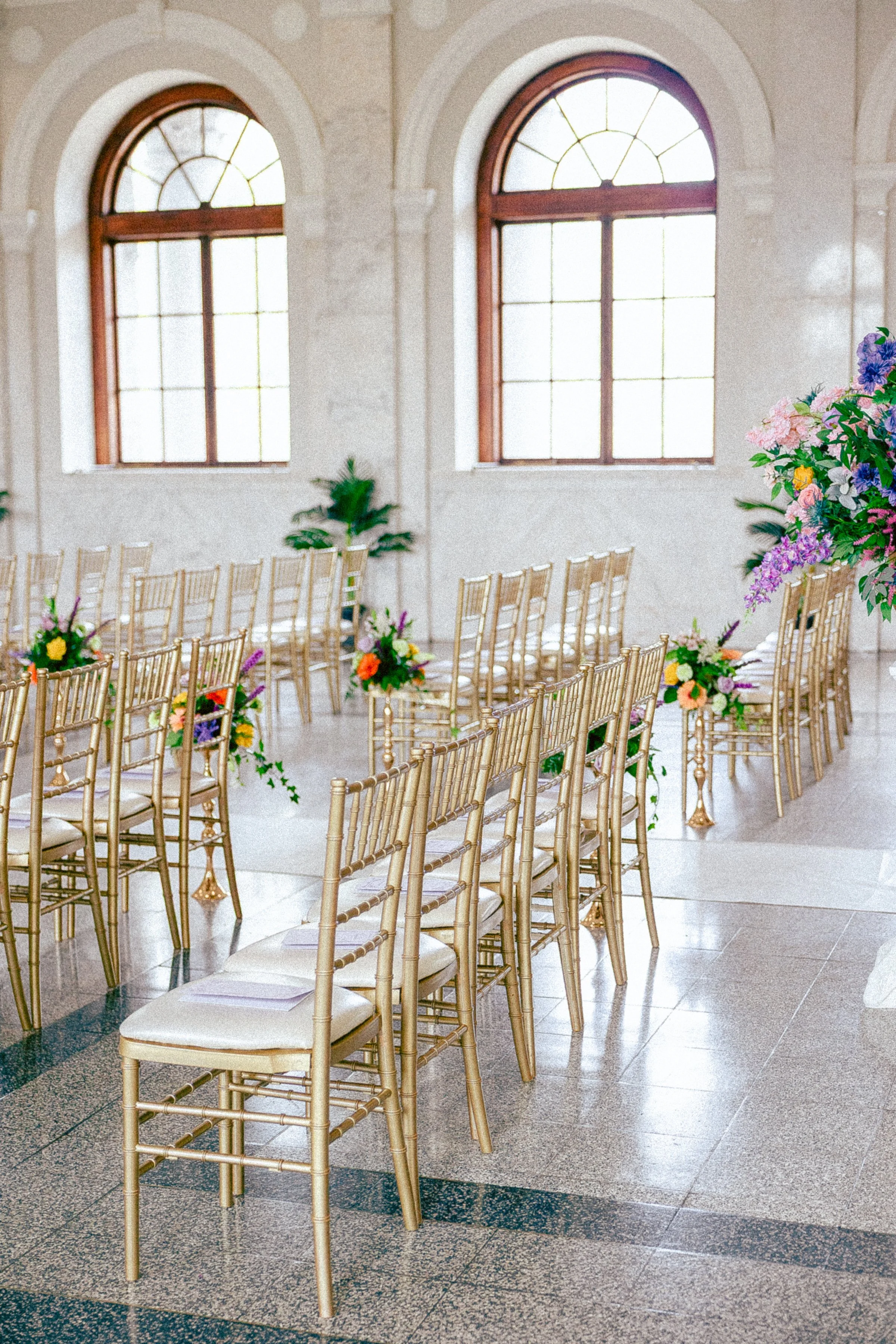 A decorated indoor venue with gold chairs arranged in rows, colorful floral arrangements at the ends of the rows, large arched windows allowing natural light, and a beige marble wall backdrop.