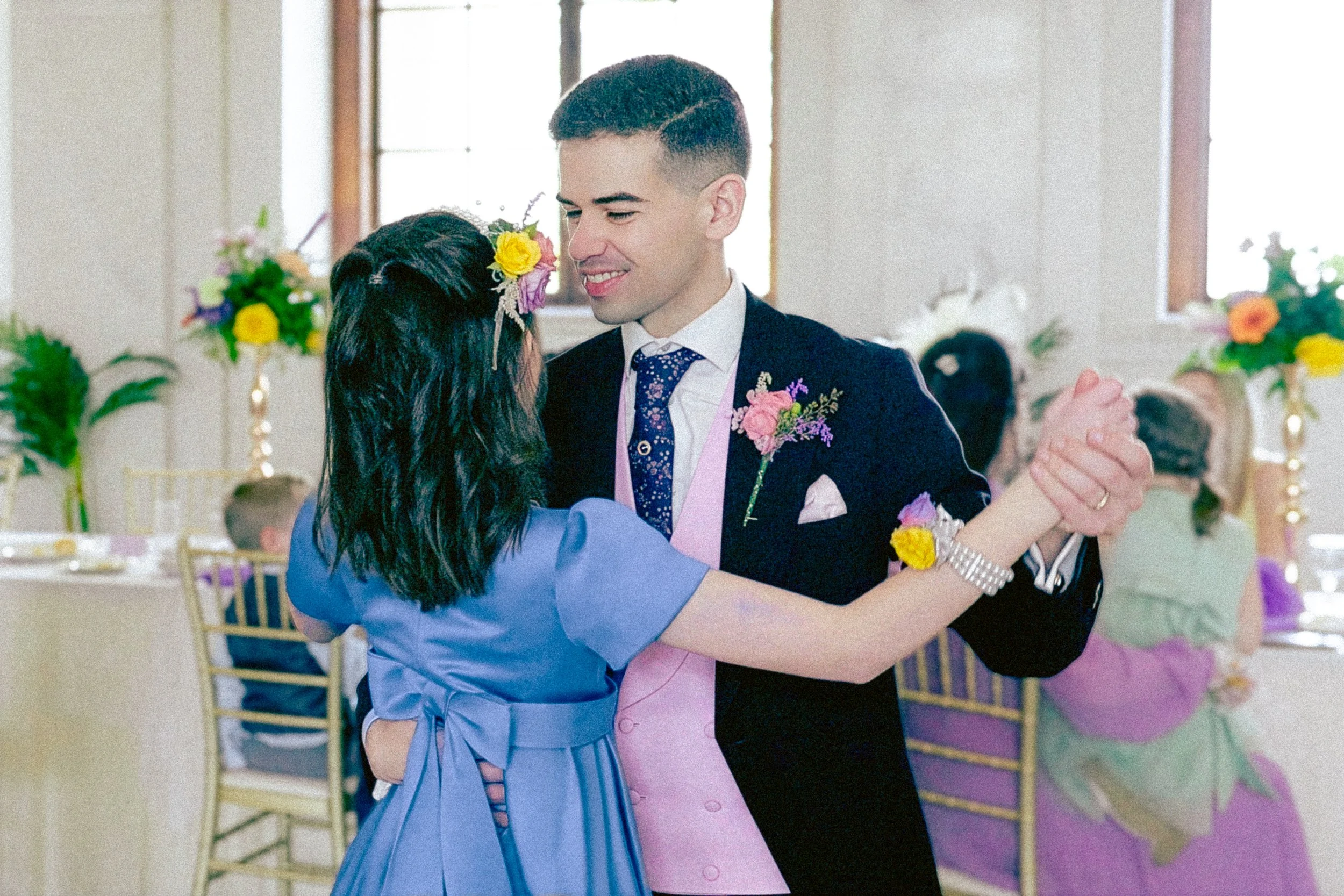 A young man and woman are dancing together at a celebration. The man is smiling and wearing a dark suit with a floral boutonniere, while the woman is in a blue dress with a floral hair accessory. They are holding hands and close to each other, surrou