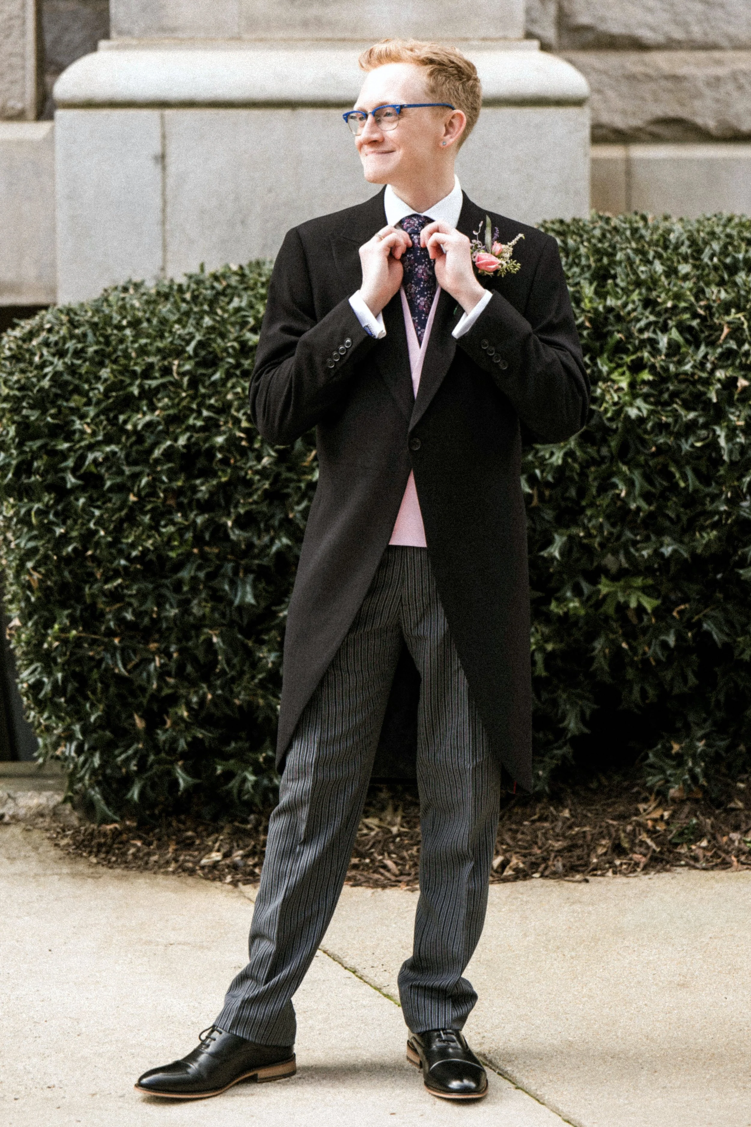 A person with short blonde hair wearing glasses, a suit, and a pink boutonniere stands outdoors on a sidewalk with greenery and stone building in the background. They are adjusting their tie and smiling.