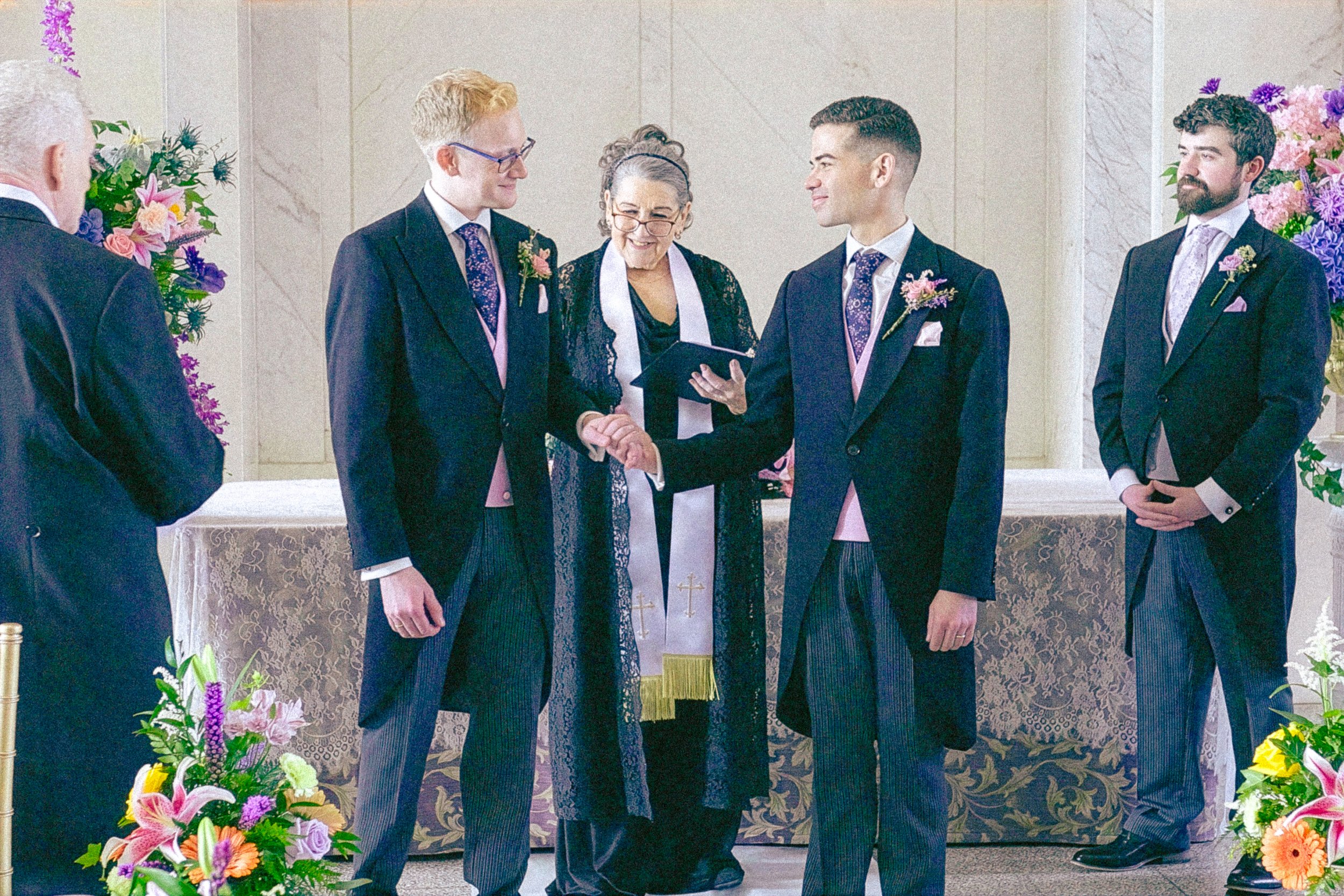 A wedding ceremony with a priest or officiant shaking hands with a groom. Other men in wedding suits are present, and there are large floral arrangements on both sides of the scene.