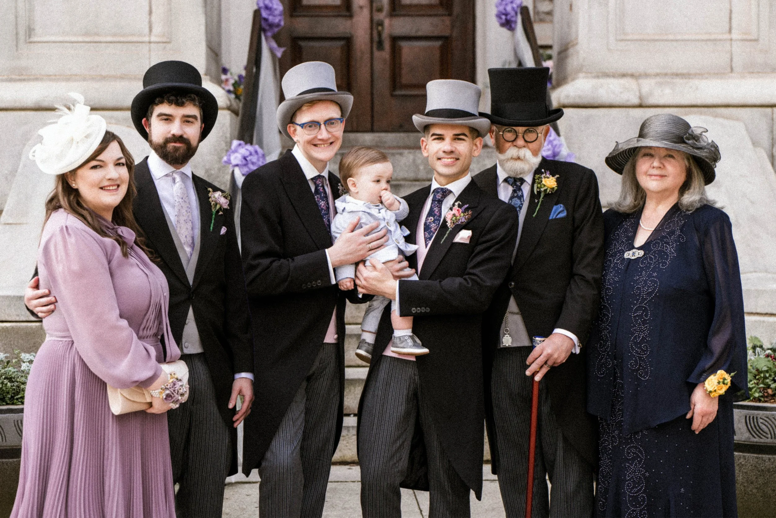 A group of seven people, including a young boy and a baby, standing on the steps of a building with stone walls. They are dressed in formal attire with vintage-style hats, and some have floral boutonnieres. The scene appears to be celebratory or comm