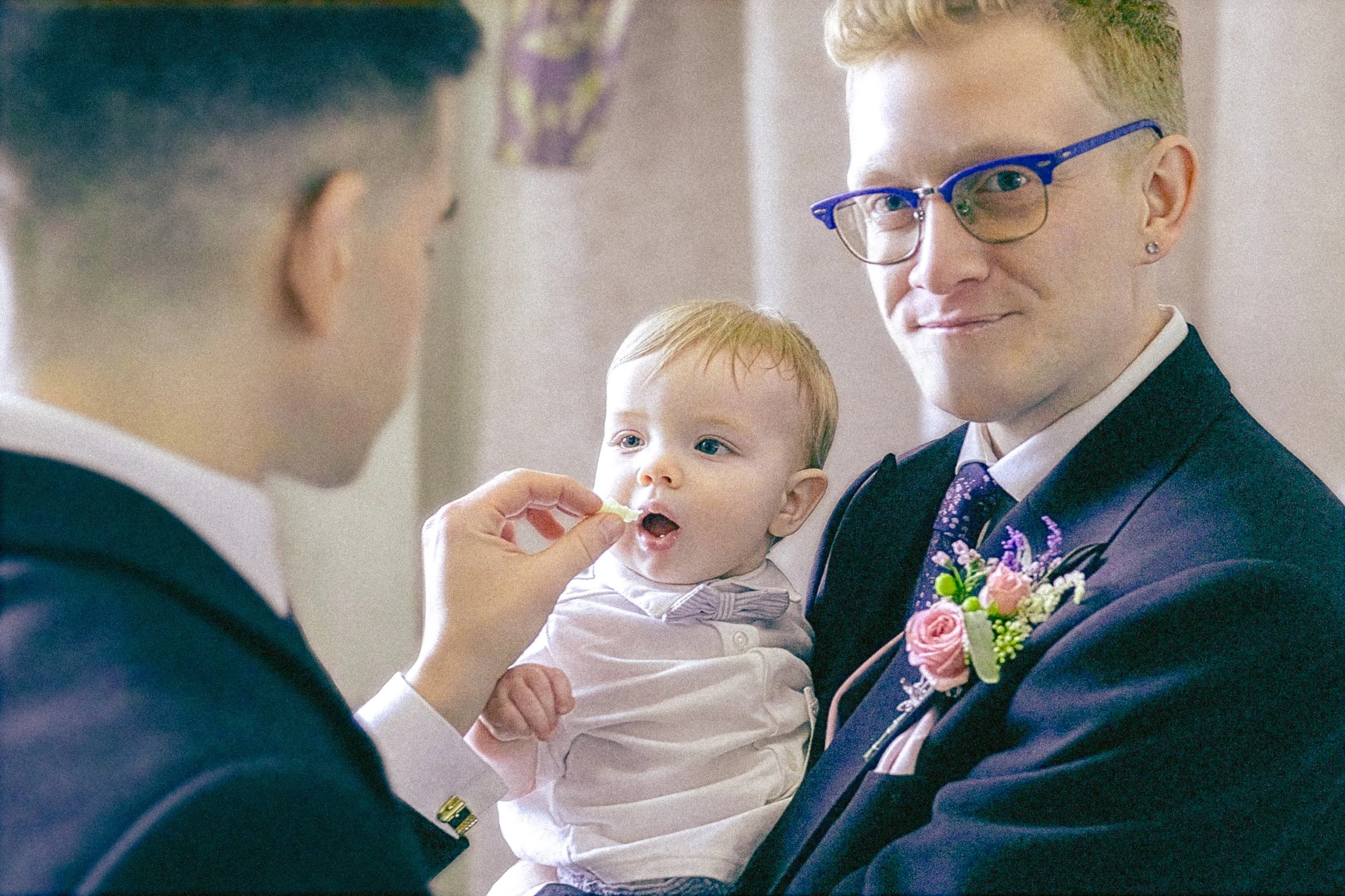 A man with blonde hair and glasses holding a young child with blonde hair, while another man, partially visible, appears to be offering the child food, at a formal event like a wedding.