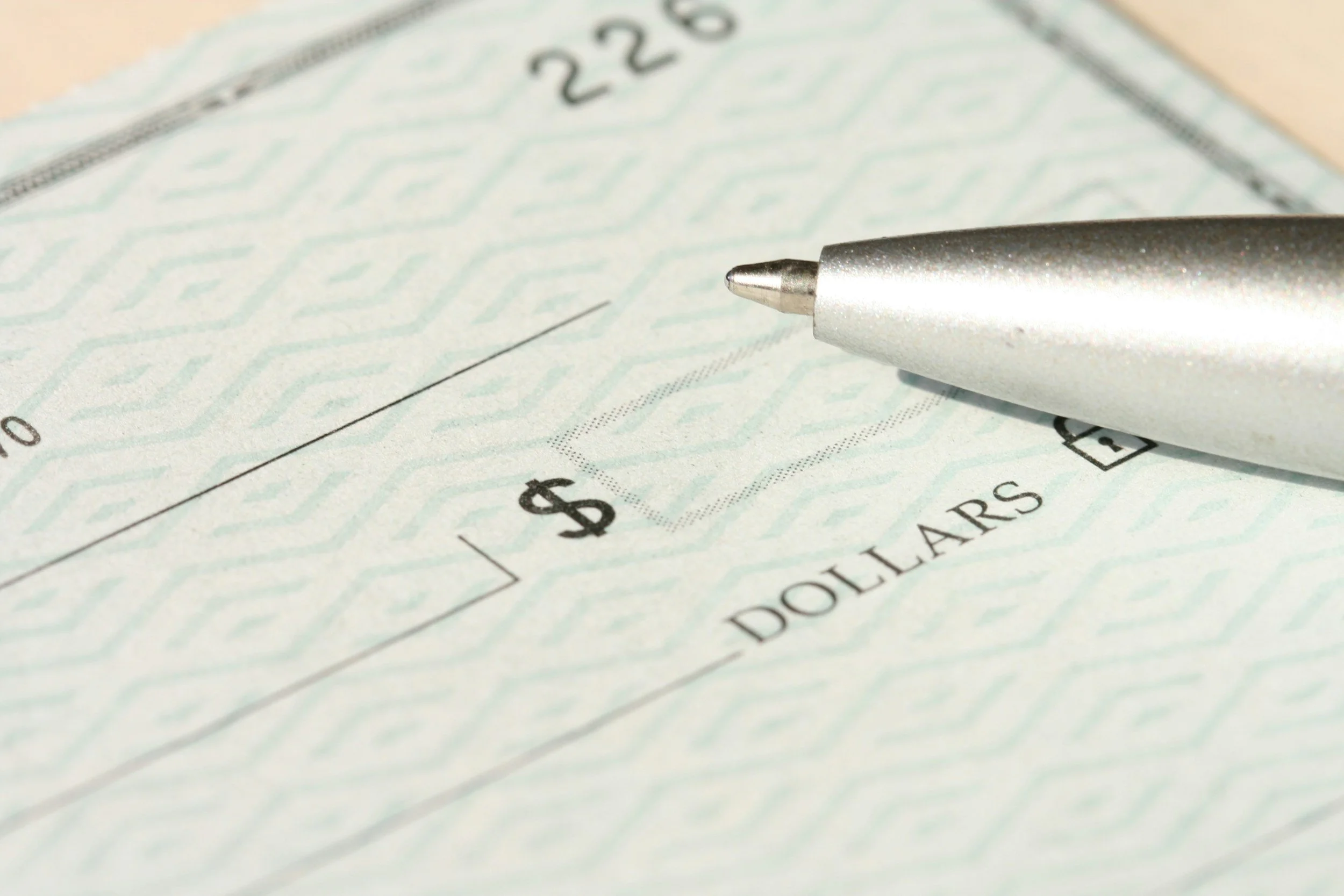 Close-up of a check with a silver pen resting on it, showing the dollar box and the word 'DOLLARS.'