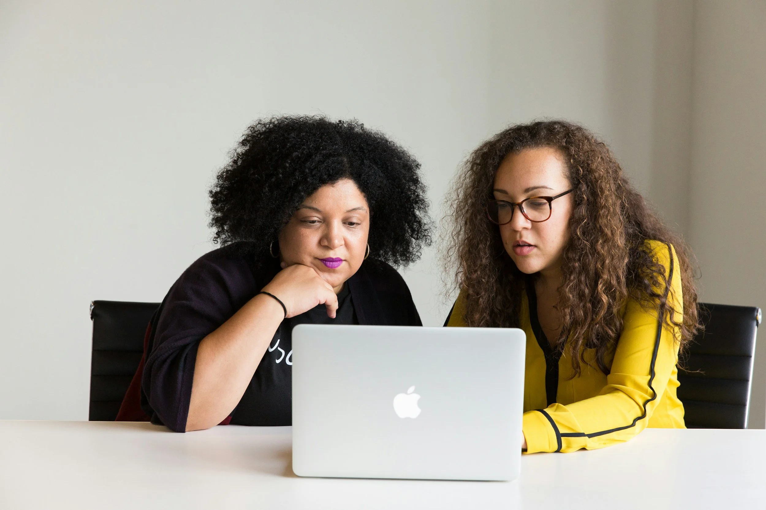 Two women looking at a silver Apple MacBook laptop on a white table in an office setting.