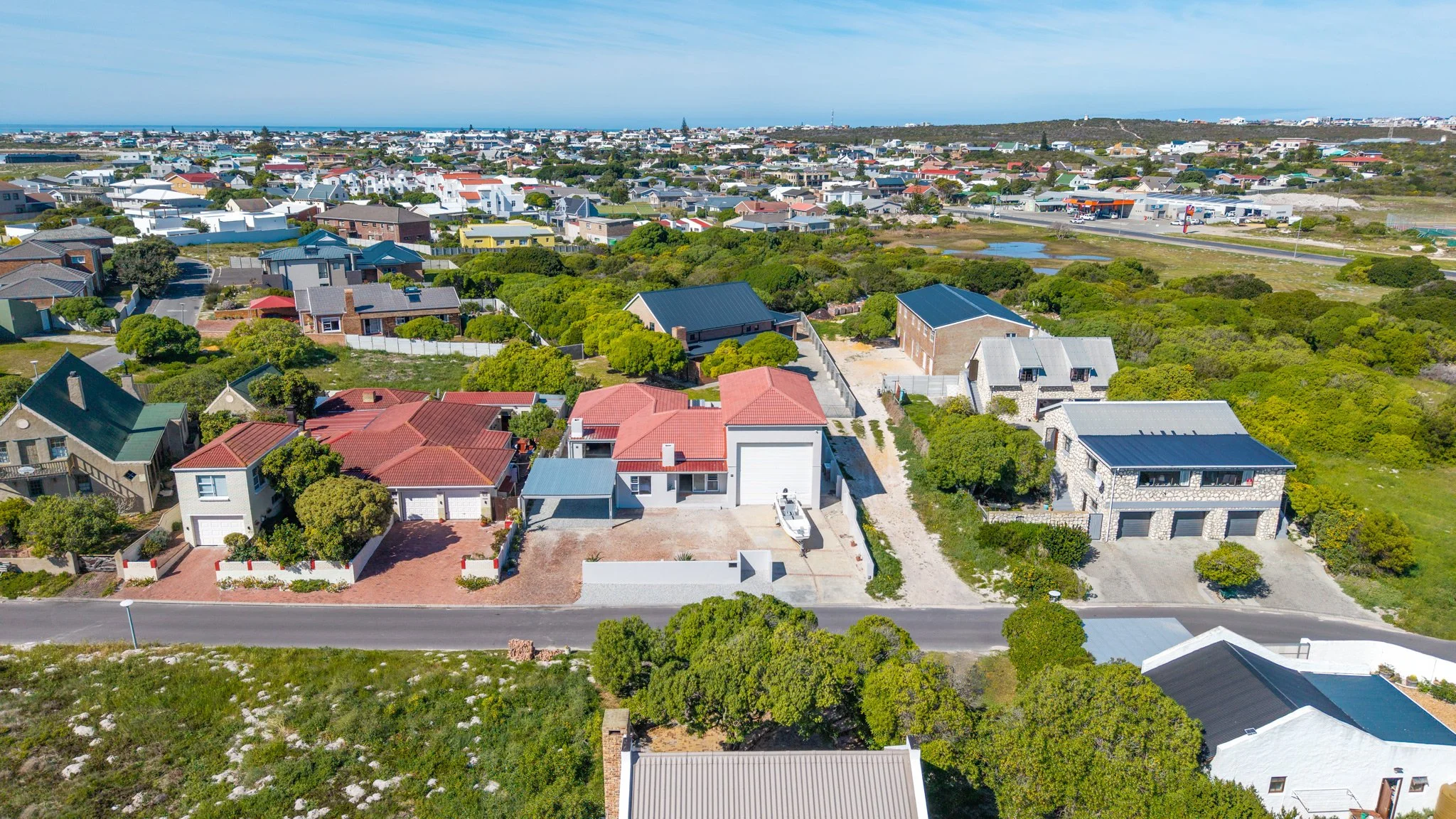 Aerial view of a neighborhood with various houses, some with red, green, and metal roofs, surrounded by greenery and trees, with a distant view of the ocean and a commercial area.
