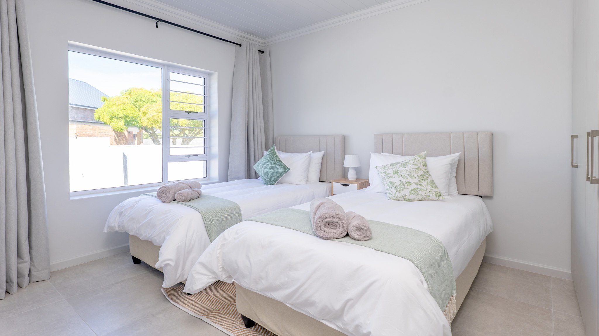 A bright bedroom with two twin beds featuring beige headboards, white bedding, green and floral pillows, and rolled towels, with a window showing trees and a building outside.