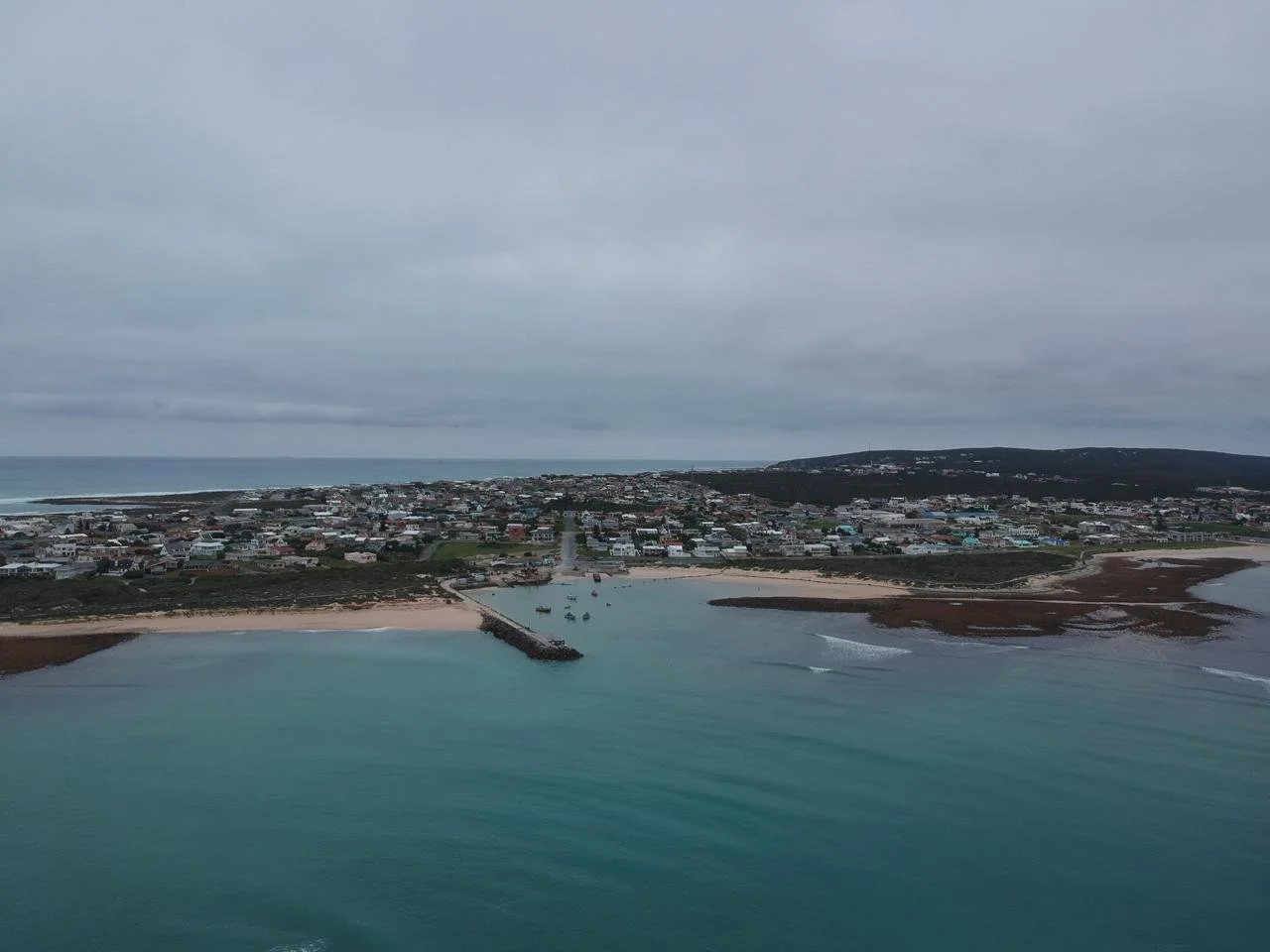 Aerial view of a coastal town with a harbor, sandy beaches, and residential buildings under a cloudy sky.