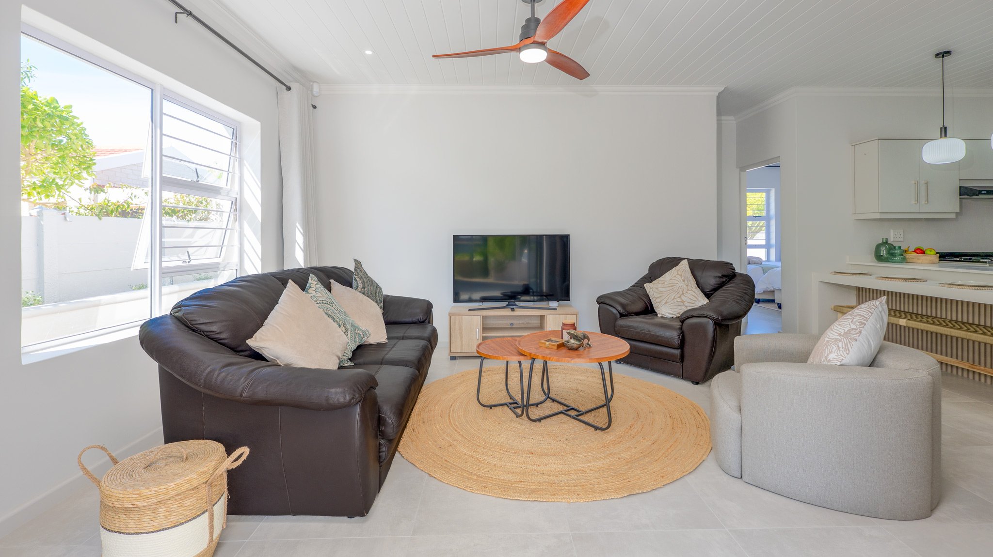 Living room with large window, black leather sofa, grey armchair, small round wooden coffee tables on a round woven rug, television on a stand, white walls, and a view into a bedroom.