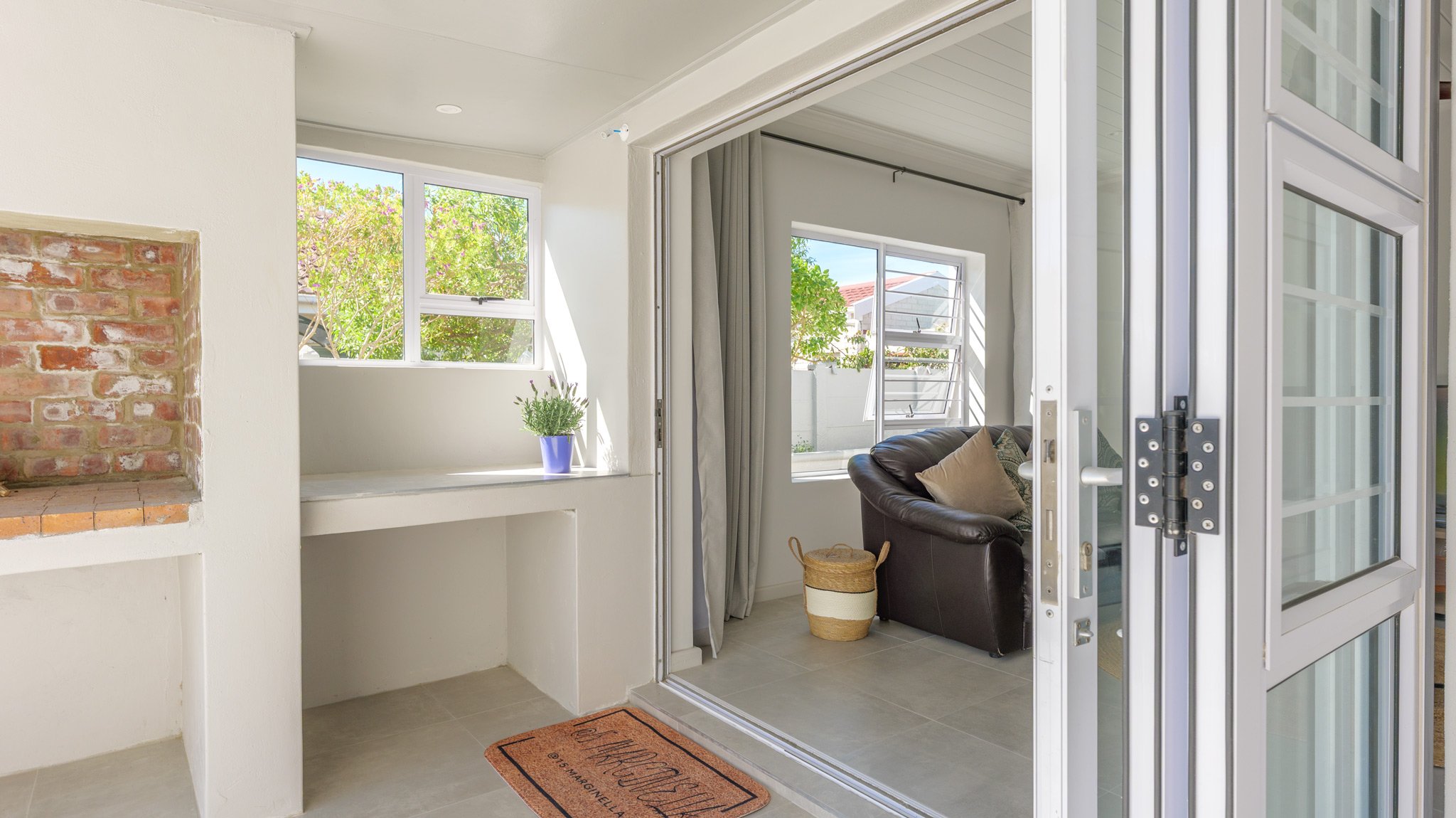 Interior view of a room with large glass windows and sliding door, beige tiled floor, and a brick fireplace corner. There is a black leather armchair with cushions, a woven basket, and a small window with a potted plant on the sill.