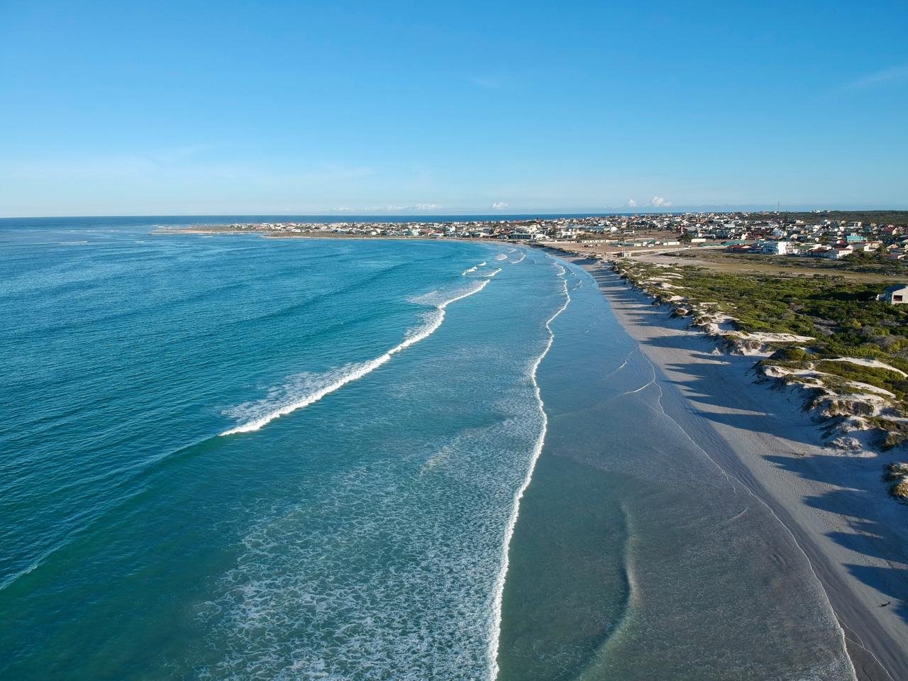Aerial view of a coastline with clear blue ocean, sandy beach, and residential area in the distance.
