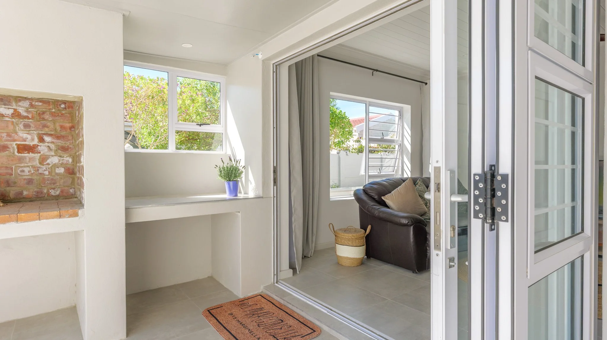 Interior of a bright living space with open sliding glass doors leading to a patio with potted plants and trees outside. The room features white walls, a brick fireplace, and a small alcove with a potted plant. Part of a brown leather sofa with pillo