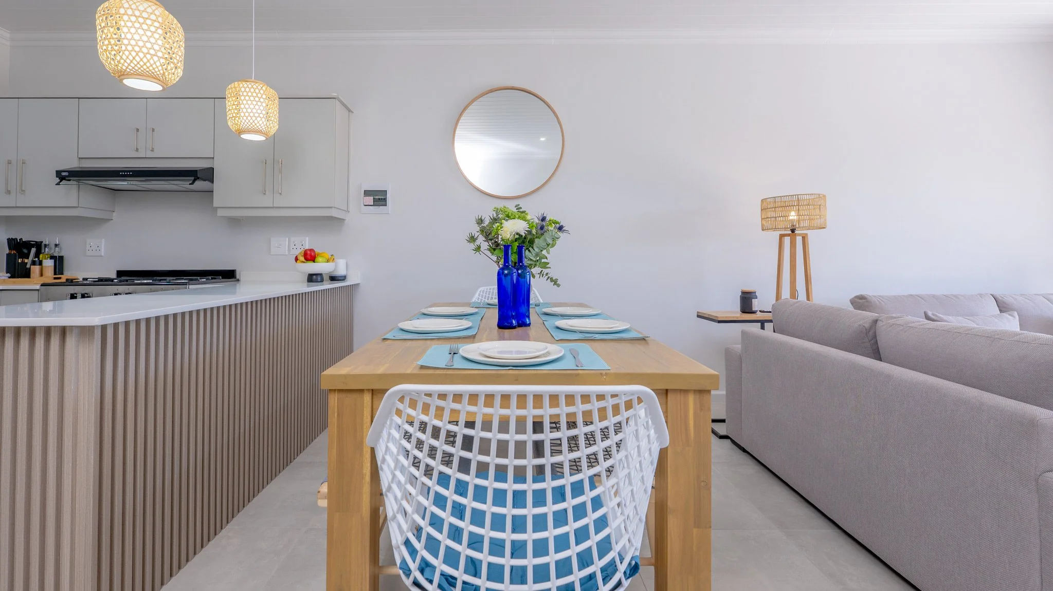 Open concept living and dining area with a wooden dining table set for six, a blue vase with flowers, a gray sofa, a wooden side table with a lamp, and a modern kitchen in the background.