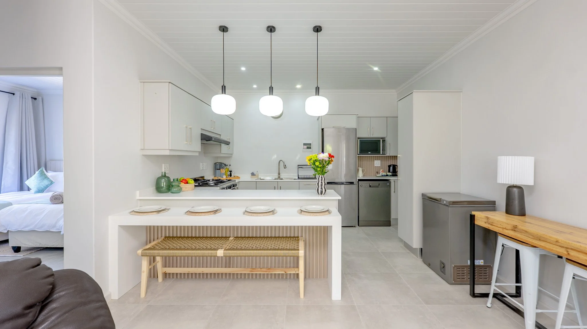 Kitchen with white cabinets, stainless steel refrigerator, and a breakfast bar with four plates and woven placemats. A vase with flowers is on the counter. Adjacent to the kitchen is a living area with a brown sofa and a side table with a lamp. In th