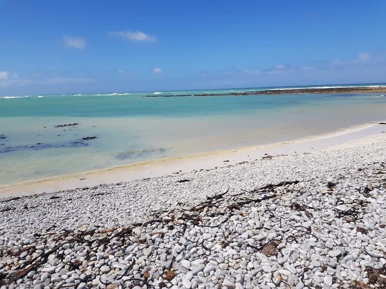 A sandy beach with white pebbles, some seaweed, and calm turquoise water under a partly cloudy blue sky.